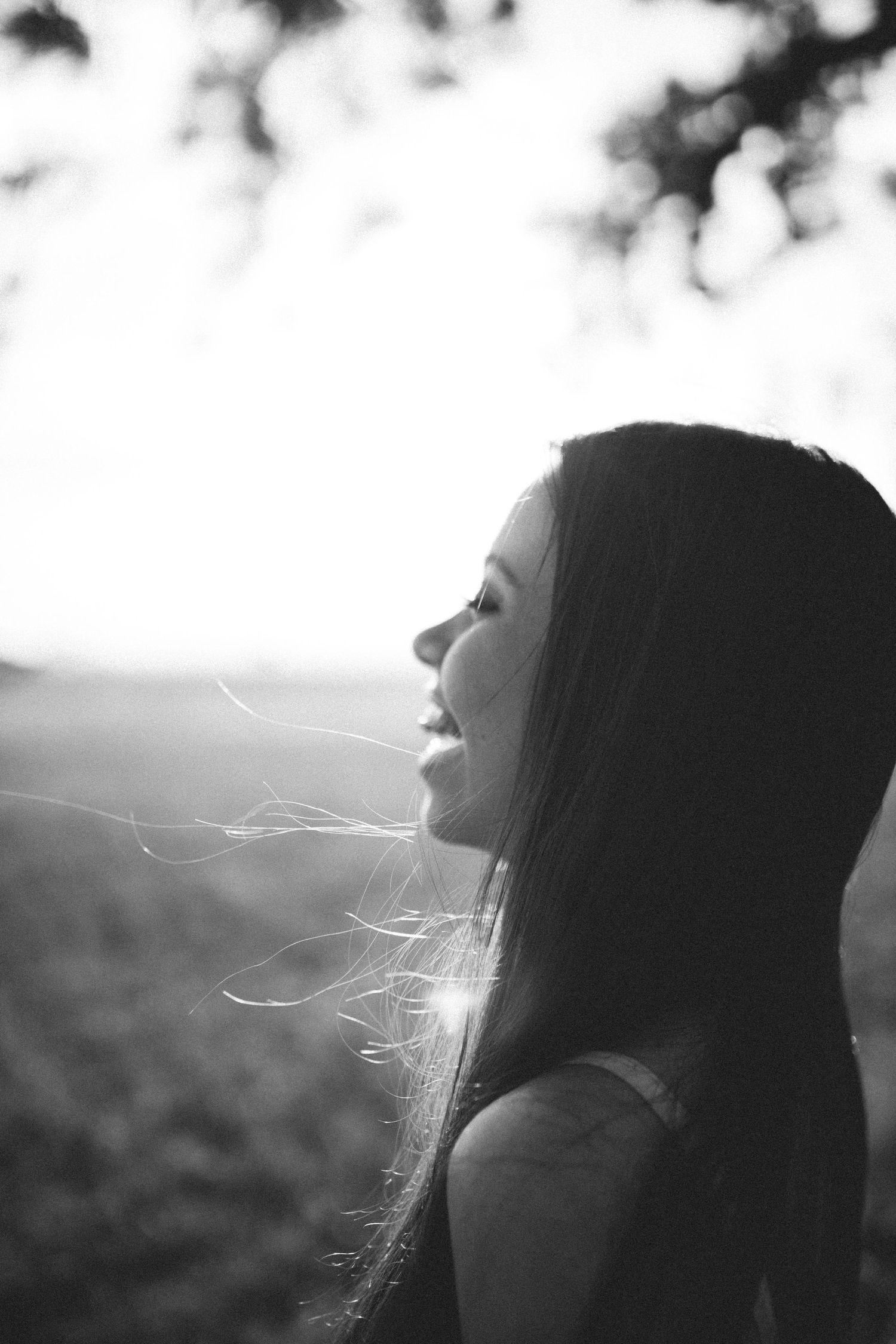 Black and white portrait of a young woman with long hair, smiling in profile against a bright backlit outdoor background.