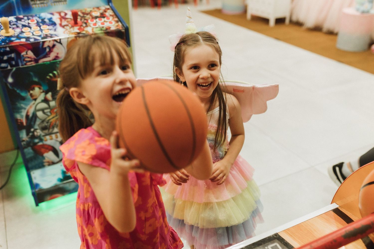 Two young girls laughing and playing with a basketball indoors, one wearing fairy wings and a colorful tutu dress.