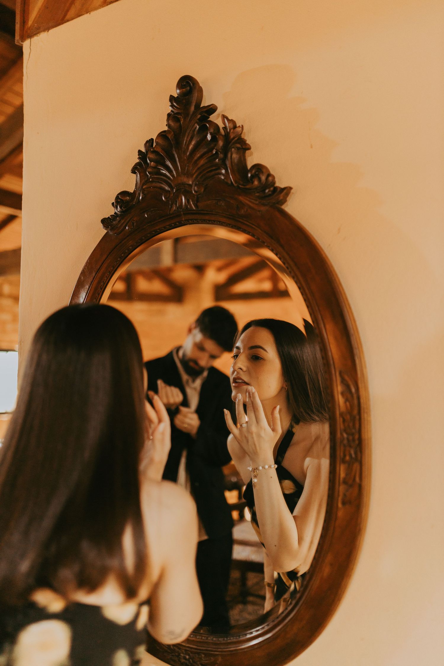 Woman applying makeup reflected in ornate vintage oval mirror with couple in background.