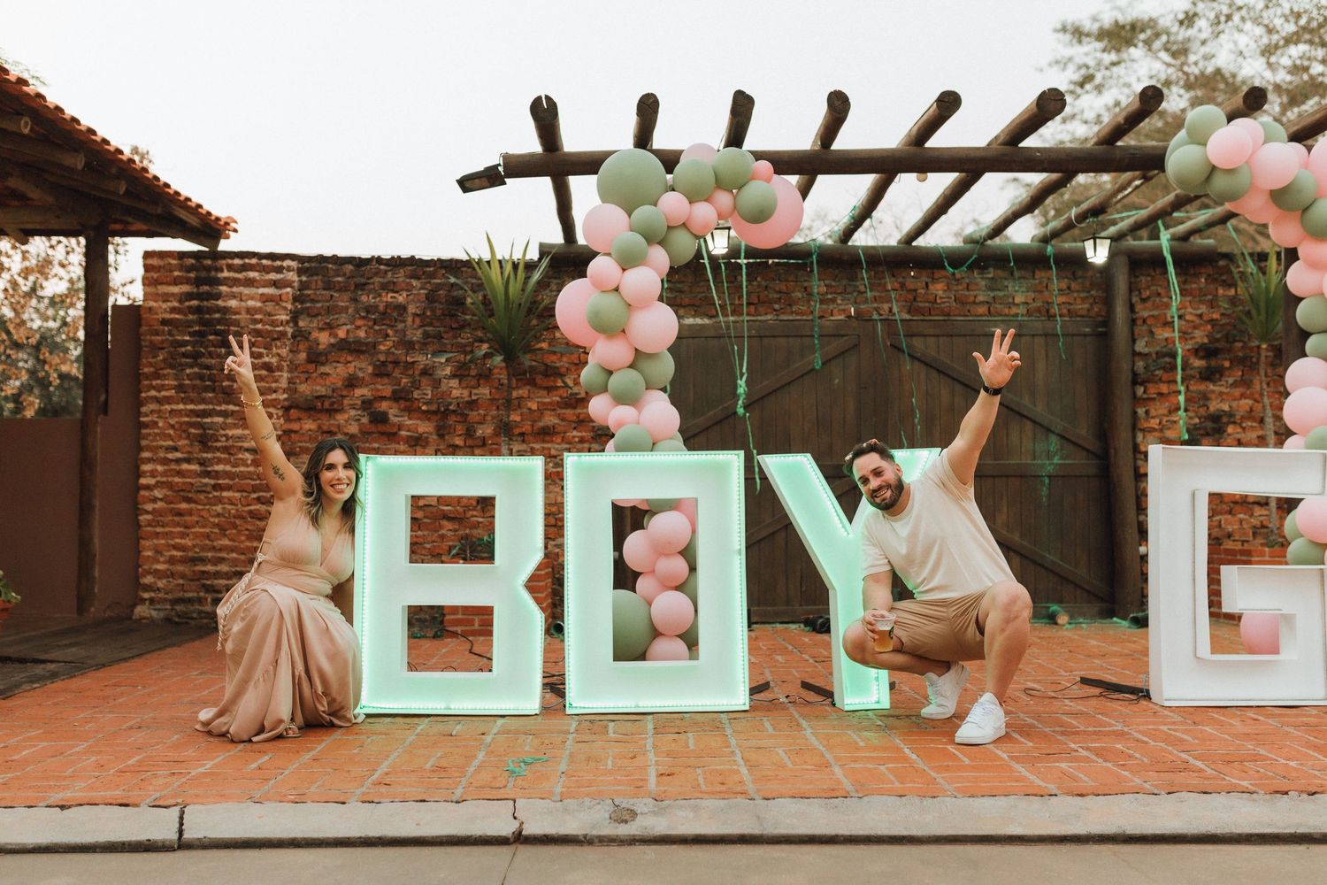 Excited couple posing beside large BOY and GIRL letters with pink and green balloons at gender reveal party.
