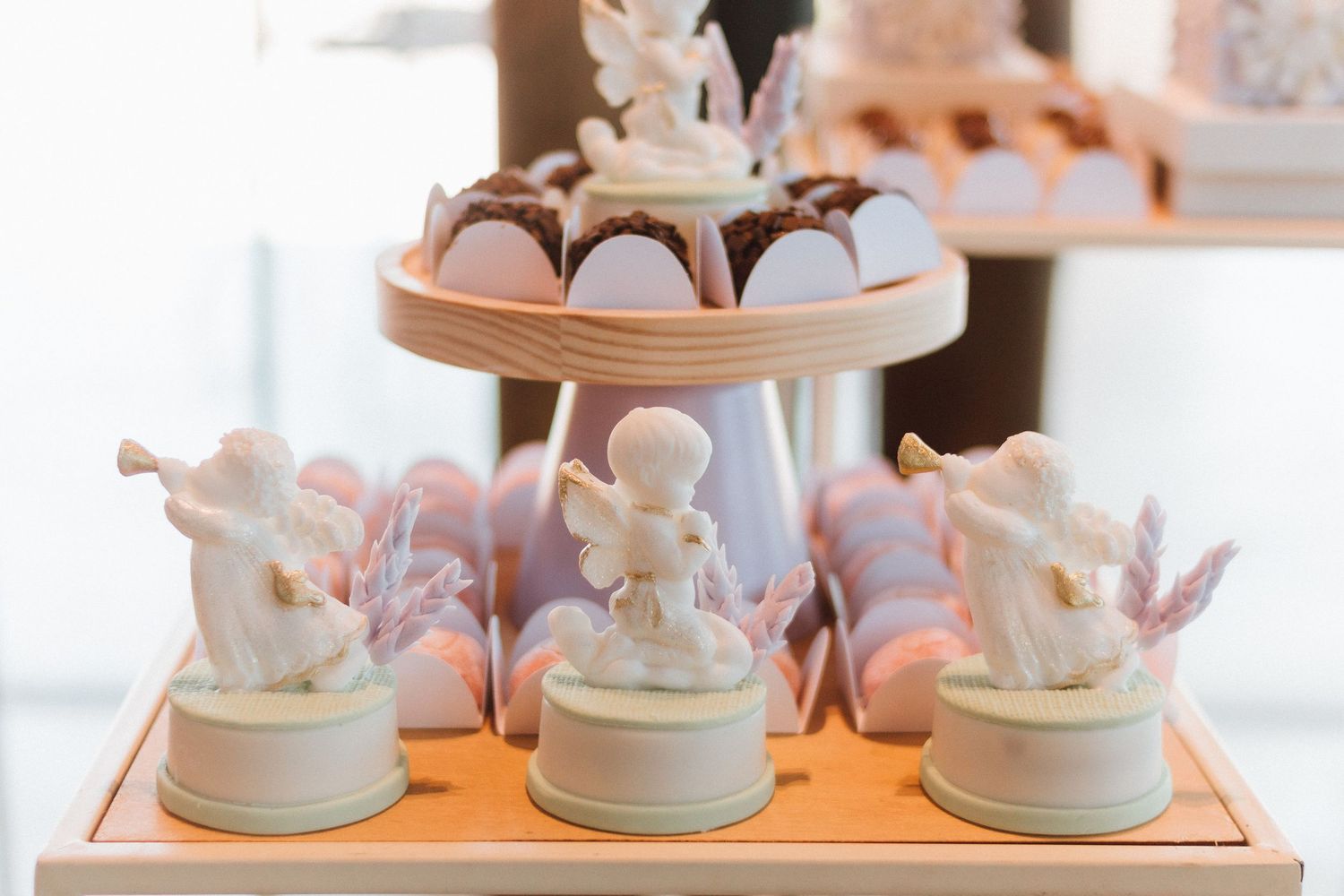 White angel figurines displayed on an orange tray beside pink macarons and chocolate cupcakes on a tiered dessert stand.