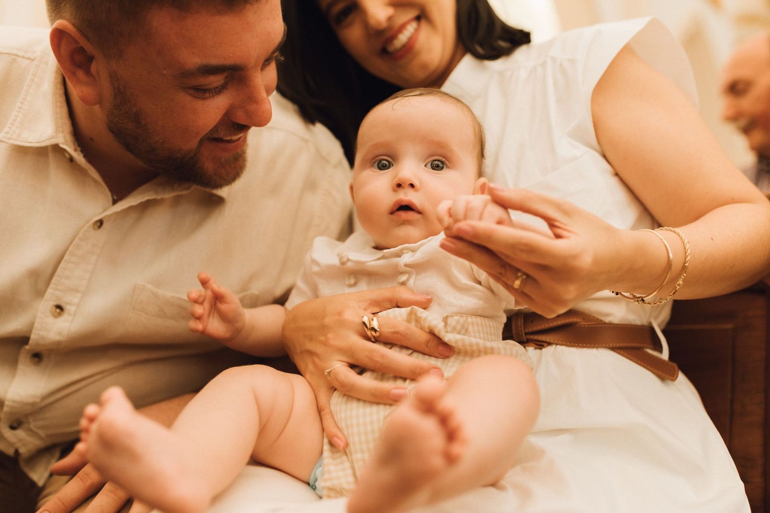 Happy parents holding their alert baby in white christening outfit during an intimate family photo session.