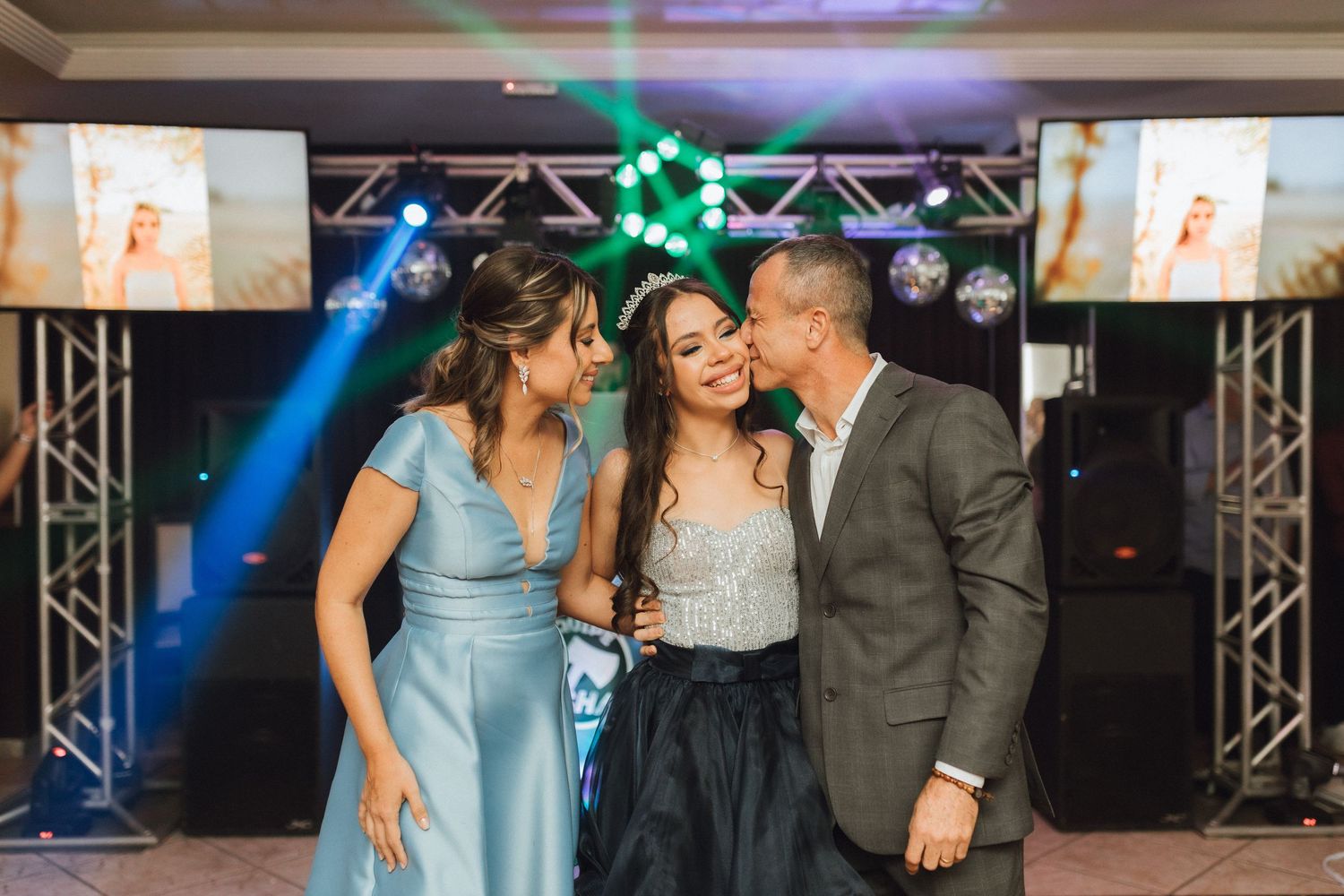 A young woman in a sparkling gown is kissed on the cheeks by her parents at a festive quinceañera celebration.
