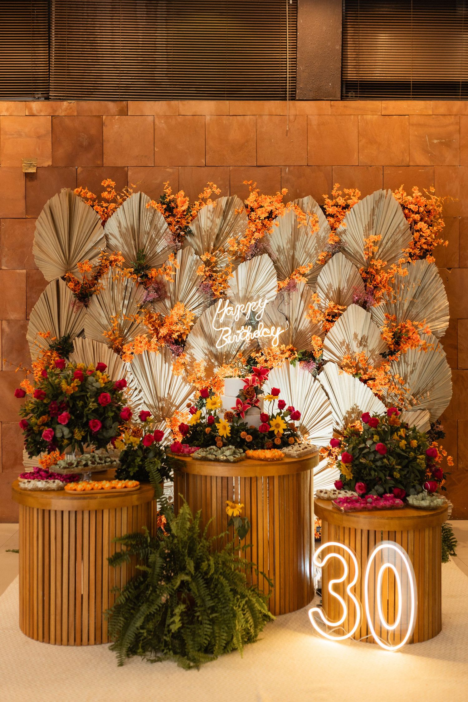 Elegant 30th birthday party decoration with floral wall, dried palm leaves, neon Happy Birthday sign, and colorful flower arrangements.