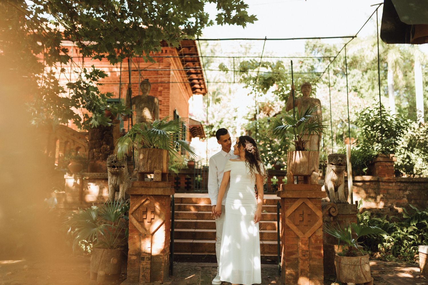 Couple in white attire standing among lush tropical plants and wooden decor at an outdoor wedding venue.