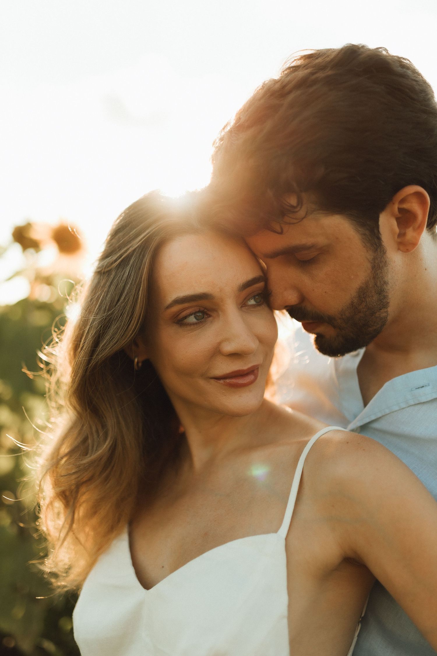 Romantic couple embracing at golden sunset, woman in white dress gazing at camera in sunflower field.