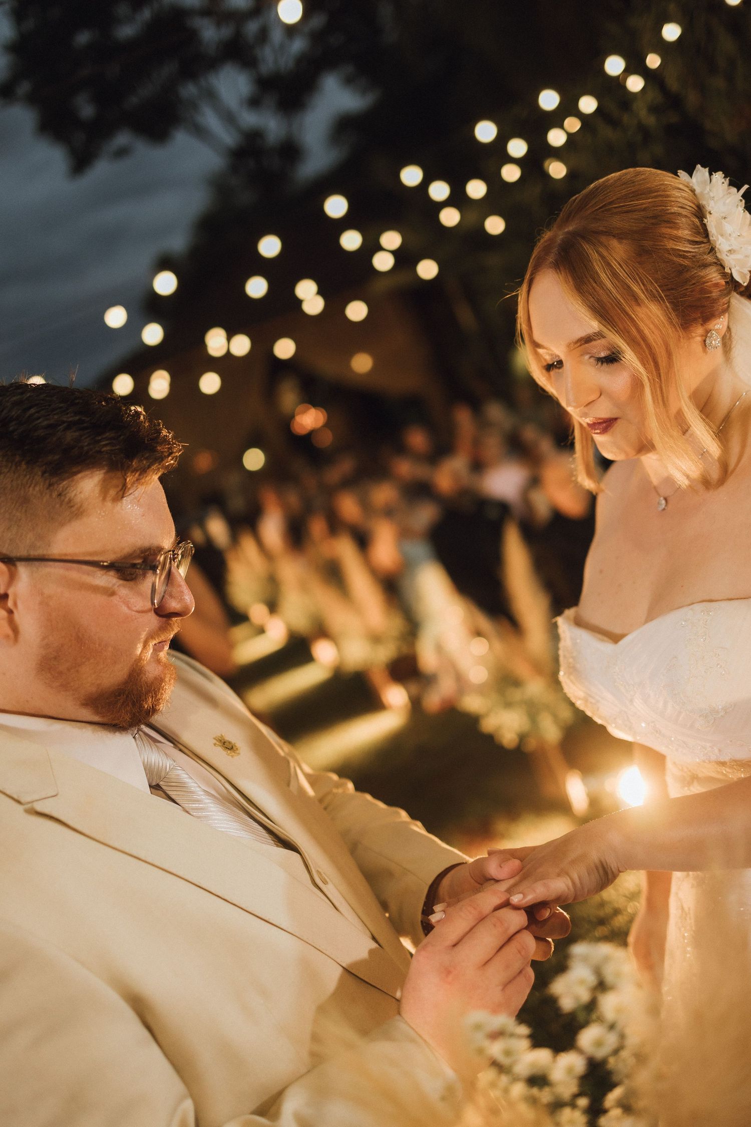 Bride places wedding ring on grooms finger during romantic outdoor evening ceremony.