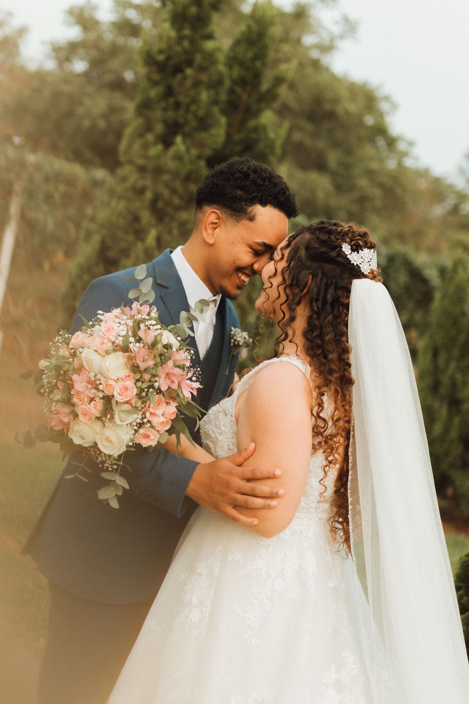 Bride and groom share a romantic forehead-touch moment, with bride holding a pink and white bouquet outdoors.