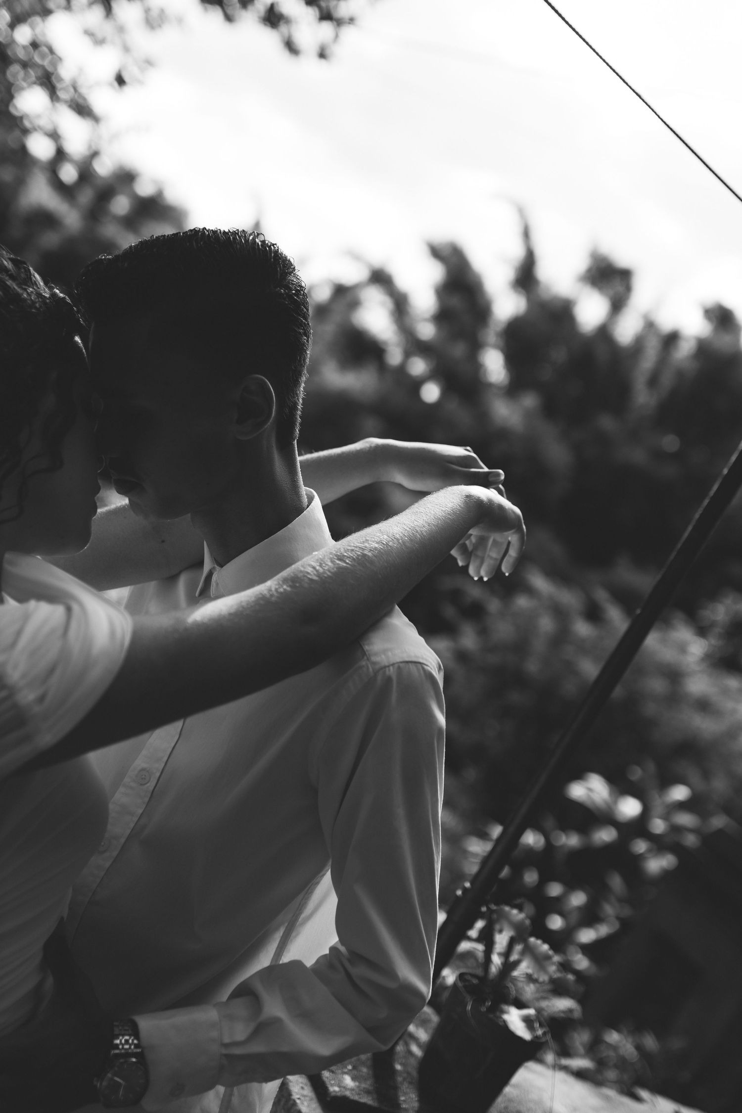 Black and white photo of a couple embracing outdoors, woman resting arm on man's shoulder near lush greenery.