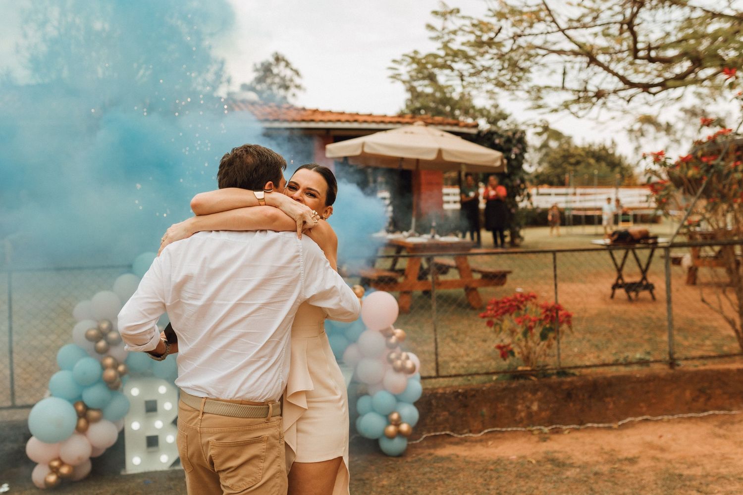 Couple embracing at a gender reveal party with blue smoke and balloon decorations outdoors.