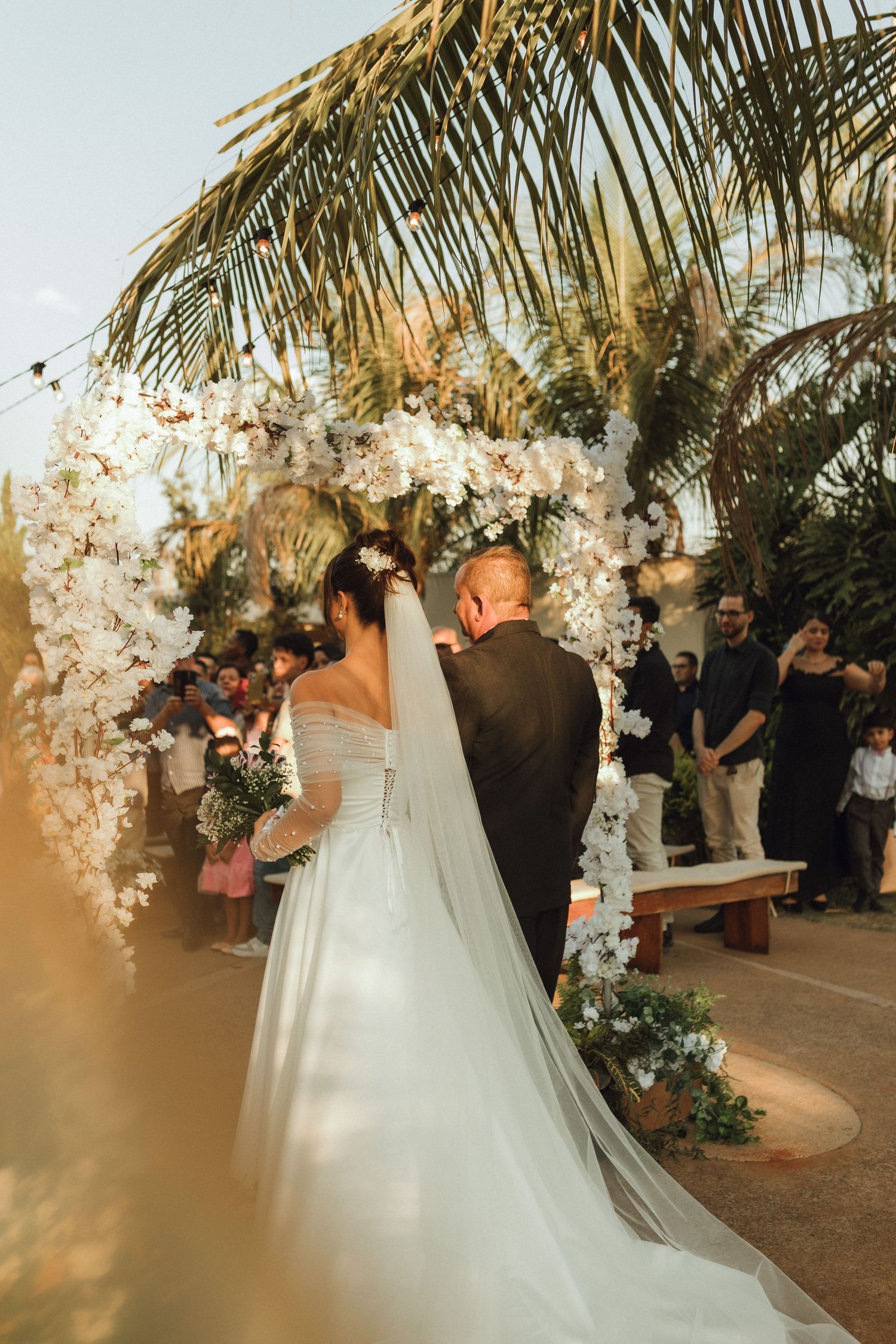 Bride and groom standing at floral arch altar during outdoor wedding ceremony at sunset.