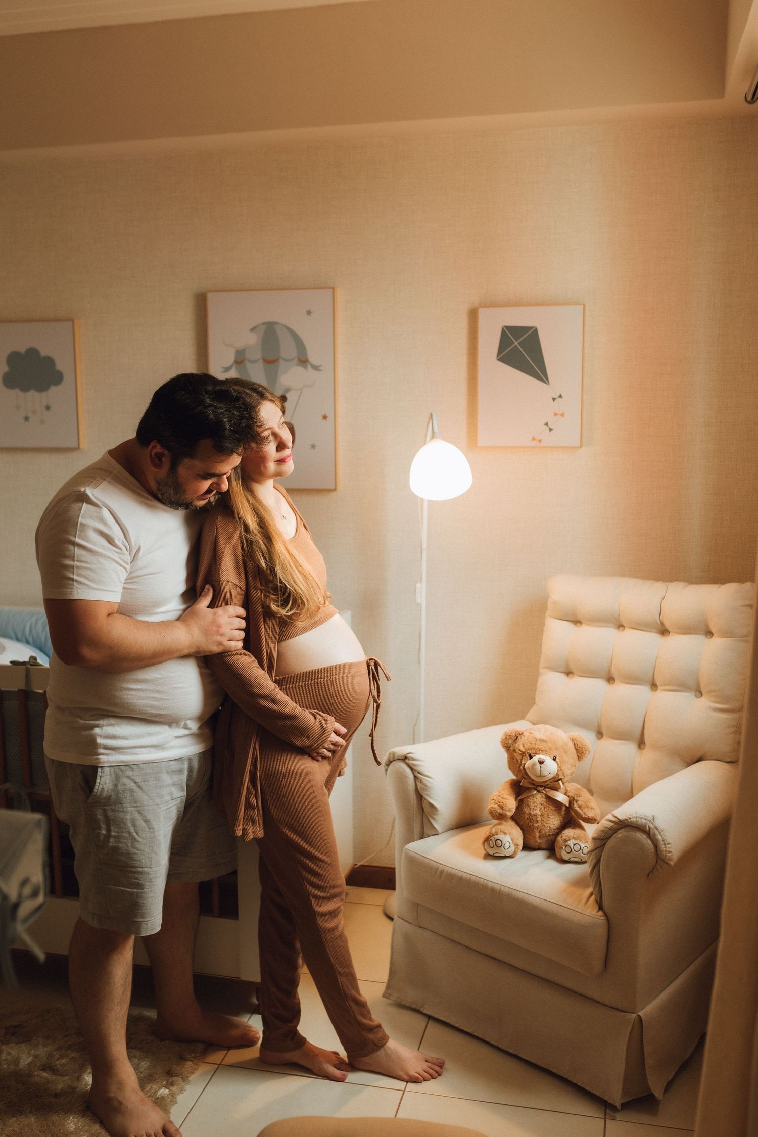Couple embraces in softly lit nursery room with teddy bear on chair nearby.