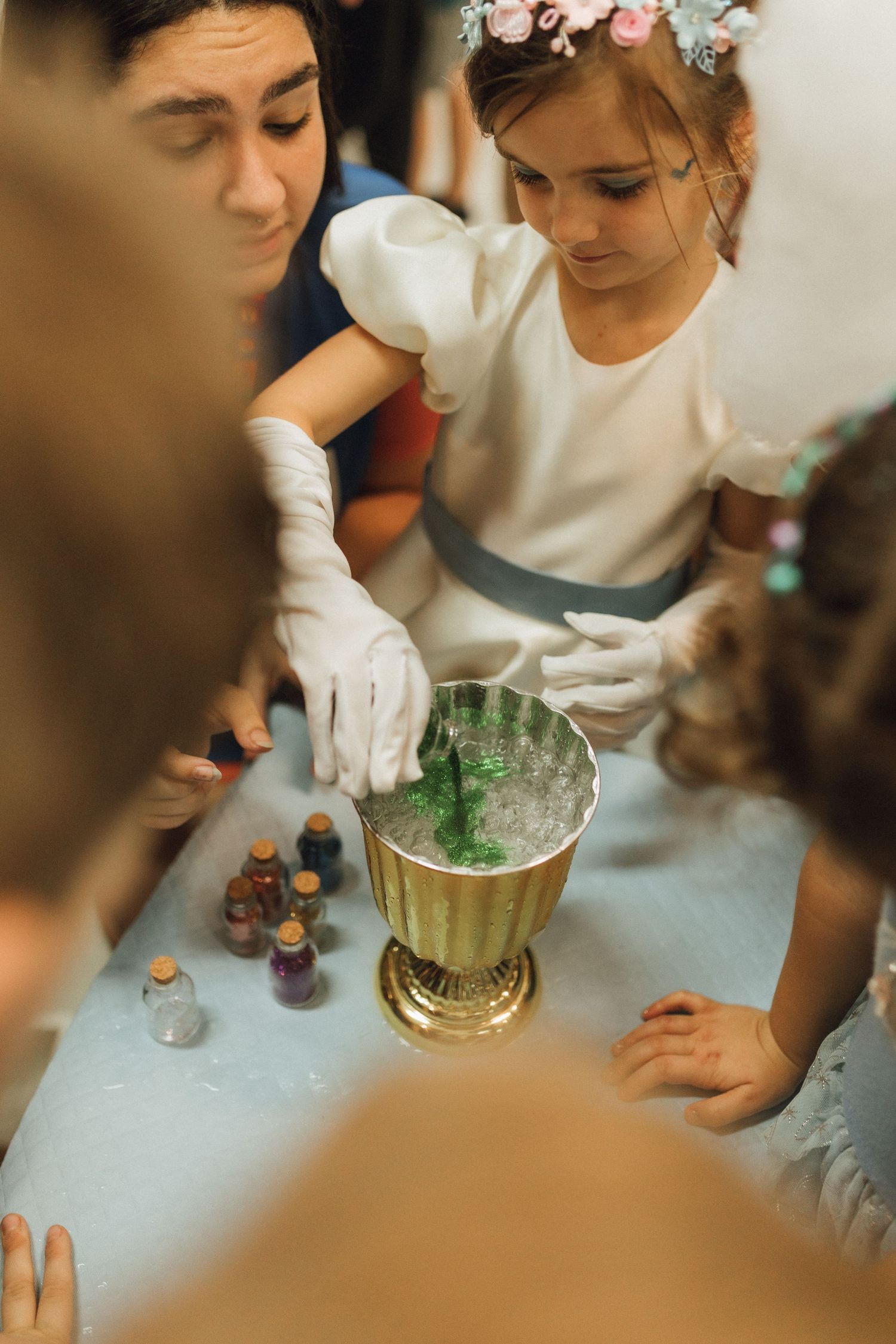 A girl in white dress and flower crown mixes green liquid in a gold bowl during a science experiment activity.