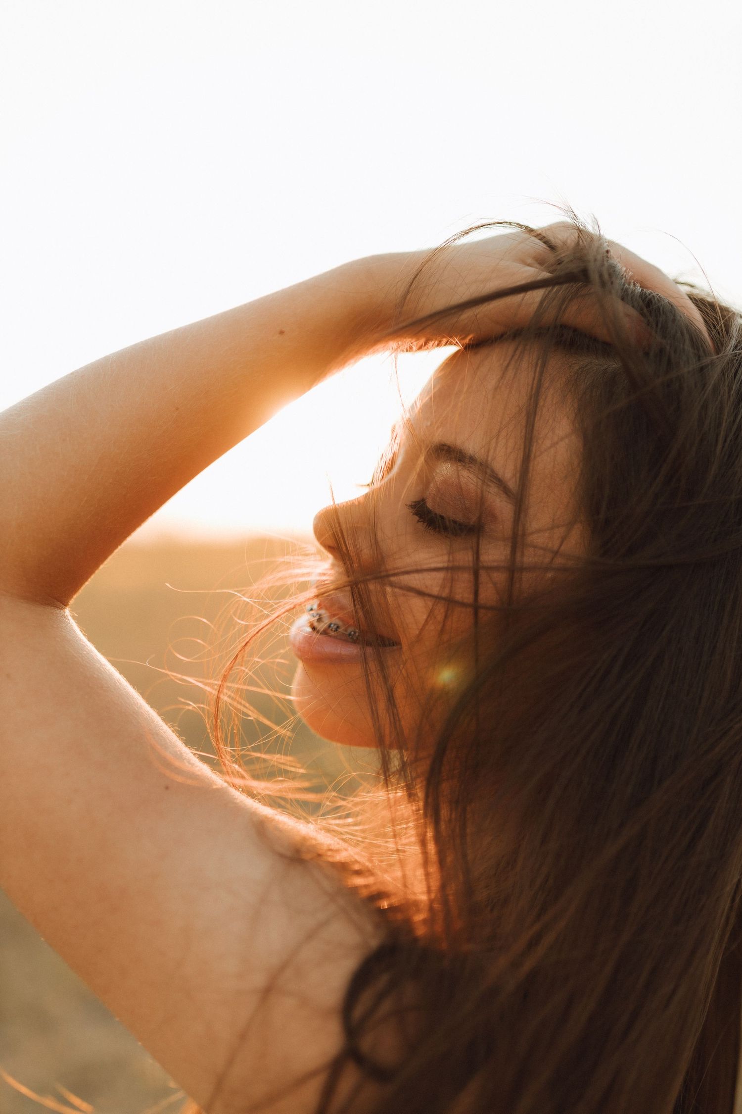 Young woman with flowing dark hair enjoys golden sunset light outdoors, eyes closed, hand in hair.