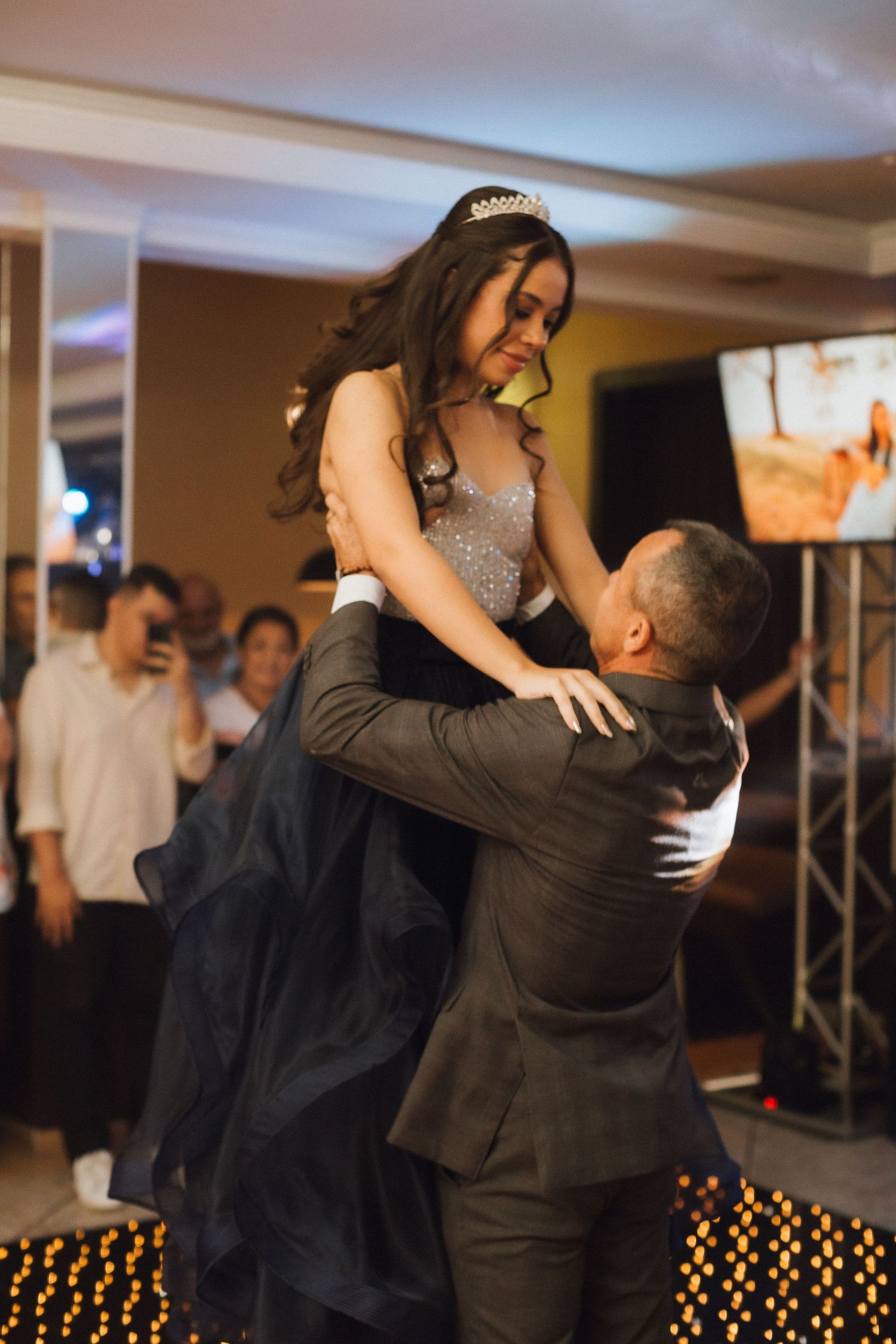 Father lifts his daughter in tiara and black ballgown during dance at her quinceañera celebration.
