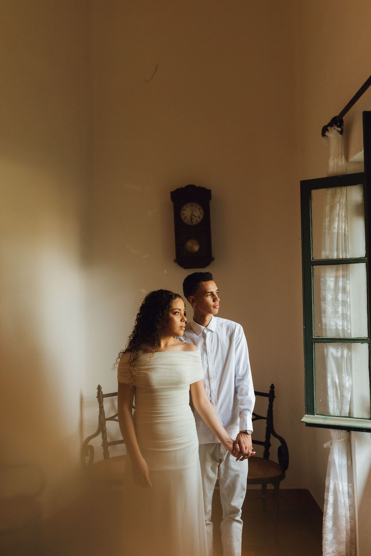 Elegant couple in white holding hands standing by window with antique clock on warm wall.