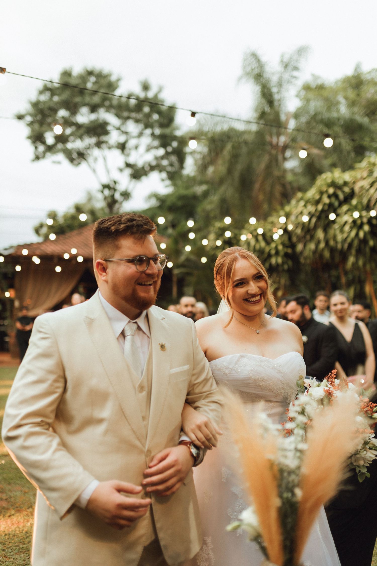 Smiling bride and groom in cream suit walk together under string lights at outdoor wedding.