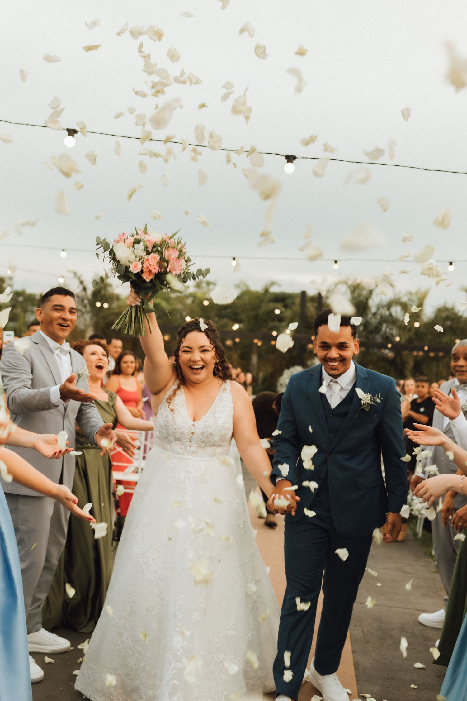 Joyful newlyweds walk down the aisle as guests toss white flower petals at their outdoor wedding reception.