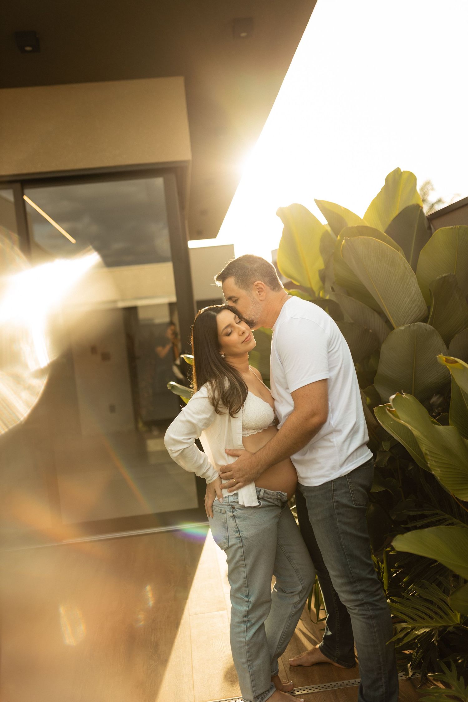 Couple embracing outdoors with pregnant belly at sunset with tropical plants background.