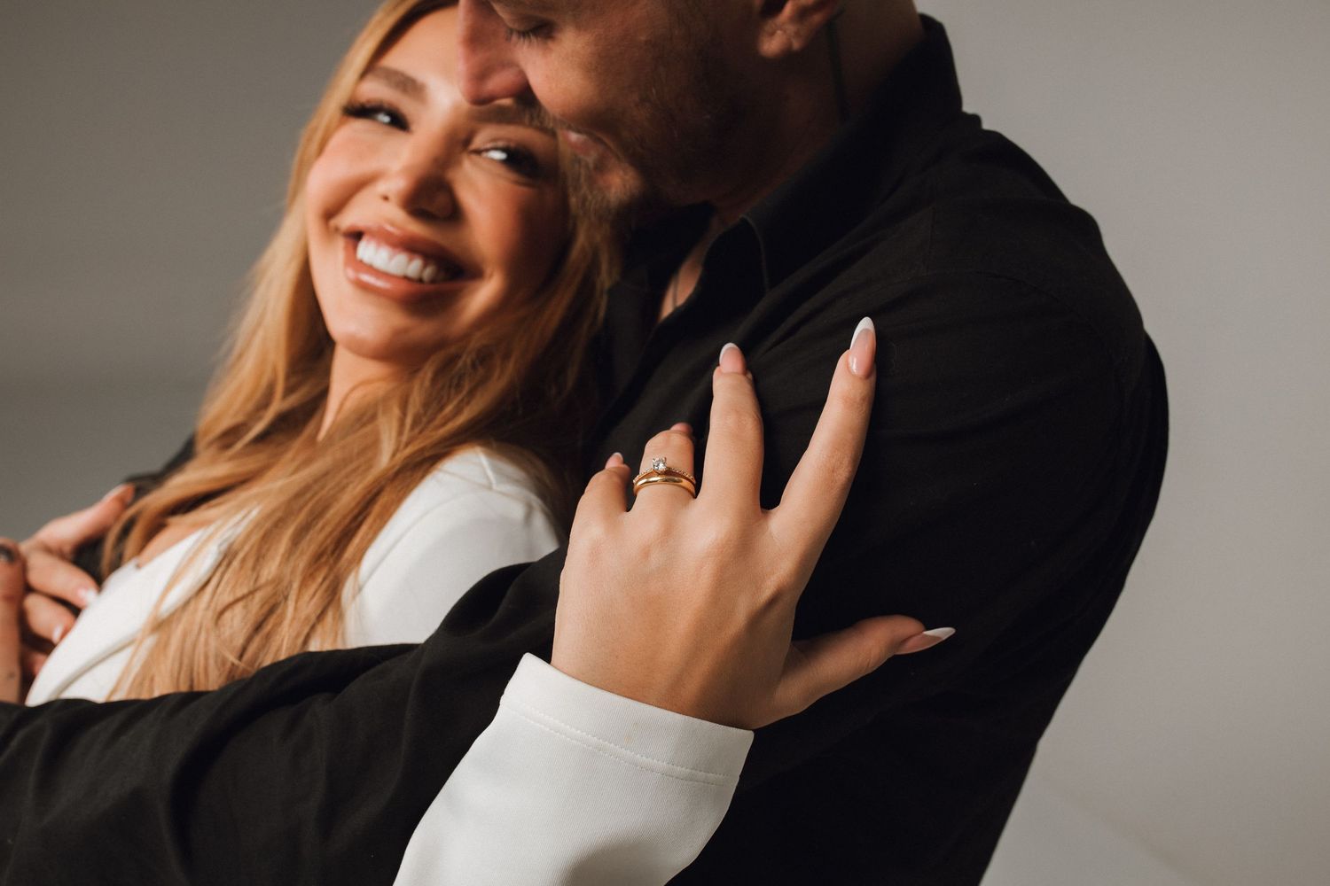 Smiling woman with blonde hair wearing engagement ring embraced by man in black jacket during couples photo shoot.