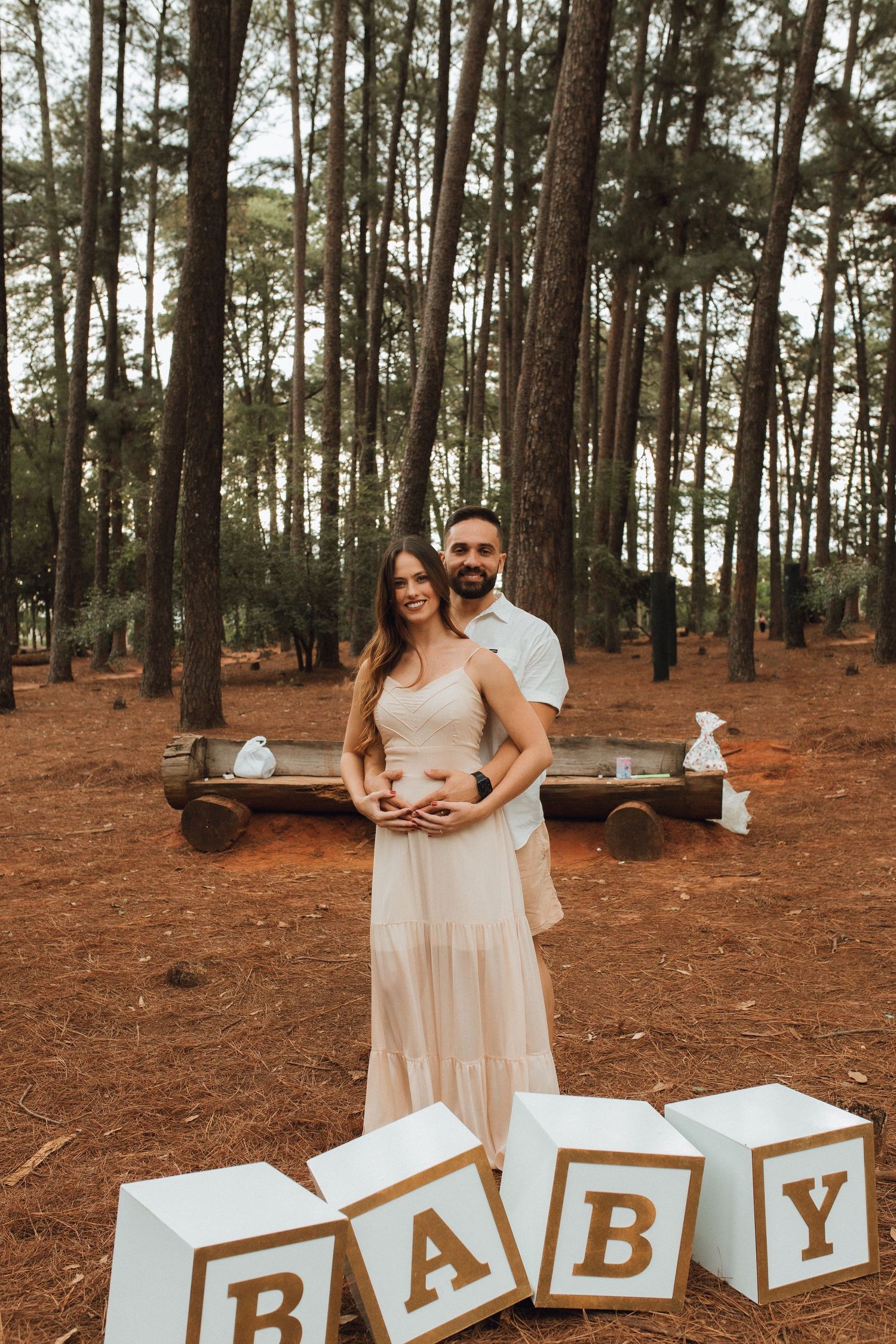Couple at outdoor baby shower with large BABY letter blocks in pine forest, pregnant woman in blush dress with partner.