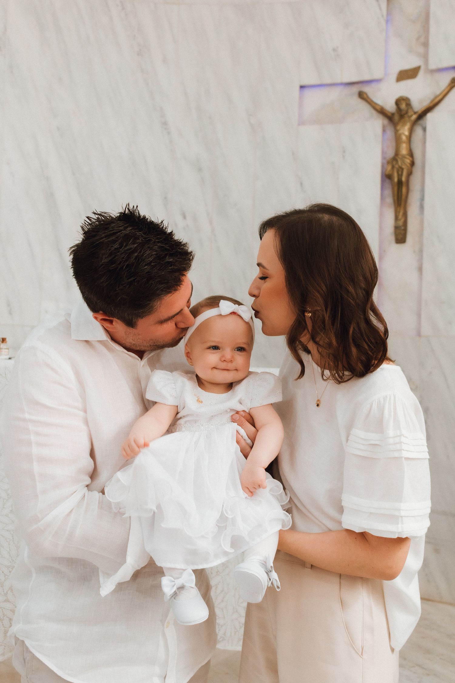 Parents kissing baby girl in white christening gown at church baptism ceremony with crucifix backdrop.
