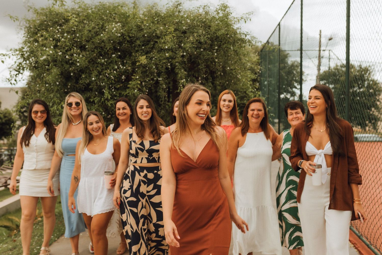 Group of women in summer dresses smiling and walking together outdoors near a tennis court and green hedges.