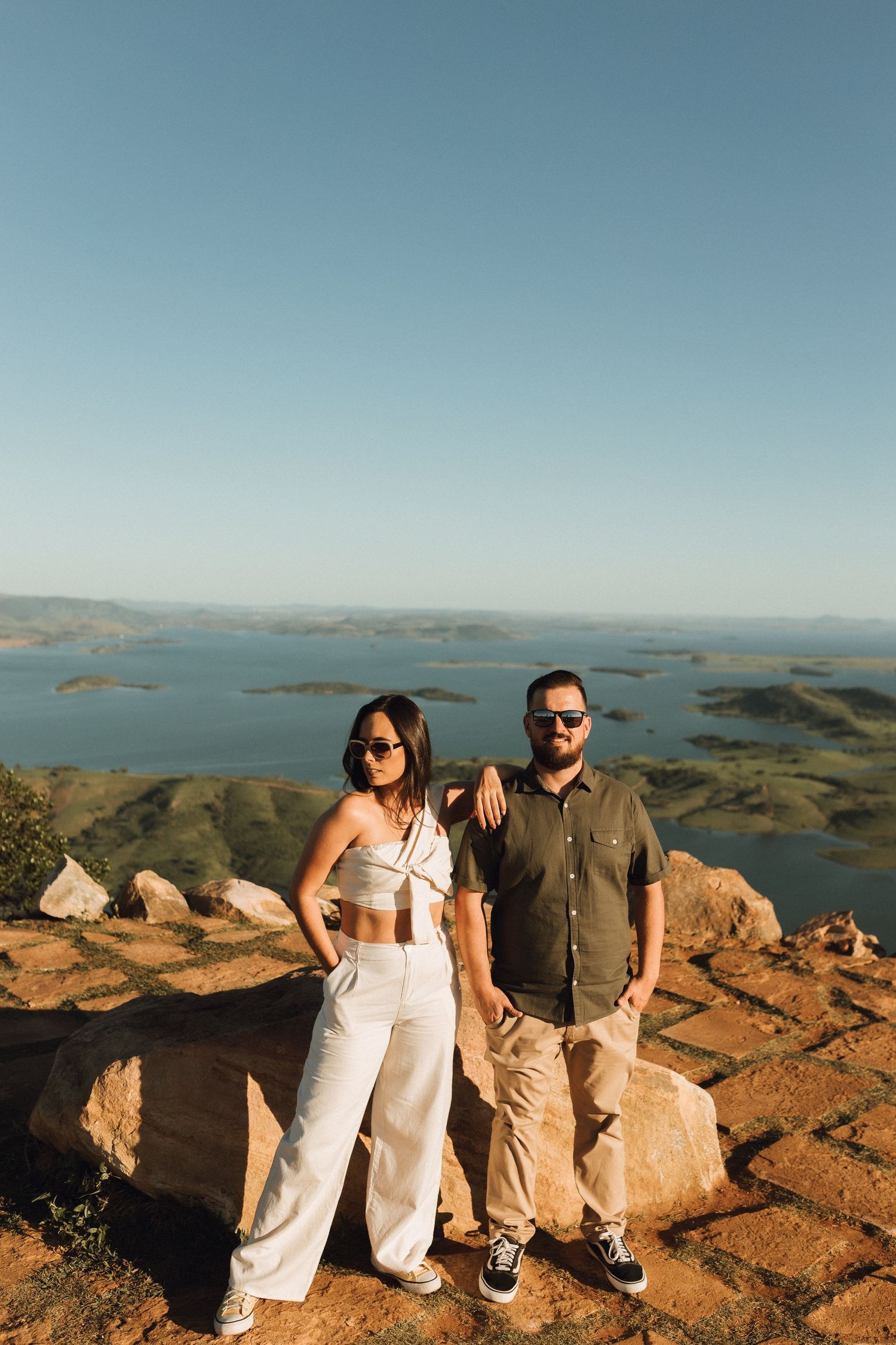 Couple posing on rocky hilltop overlook with stunning lake and island views under clear blue sky.