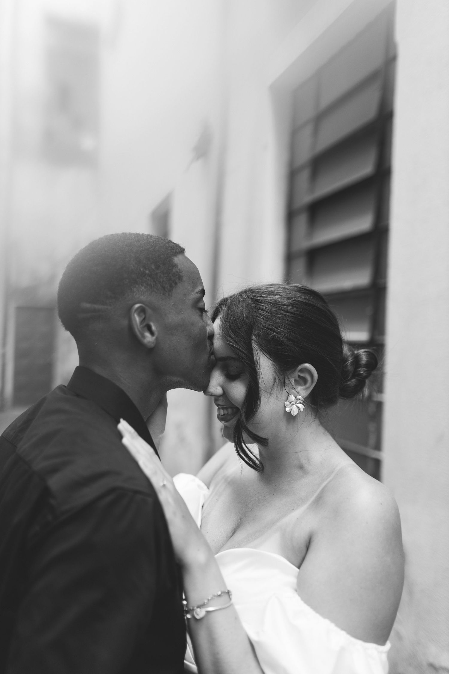 Black and white photo of a groom kissing his smiling bride on the cheek outdoors near a building wall.