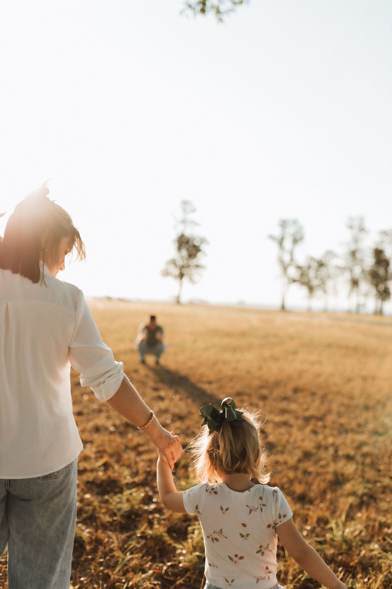 Mother and young daughter holding hands walking through a sunlit golden field.