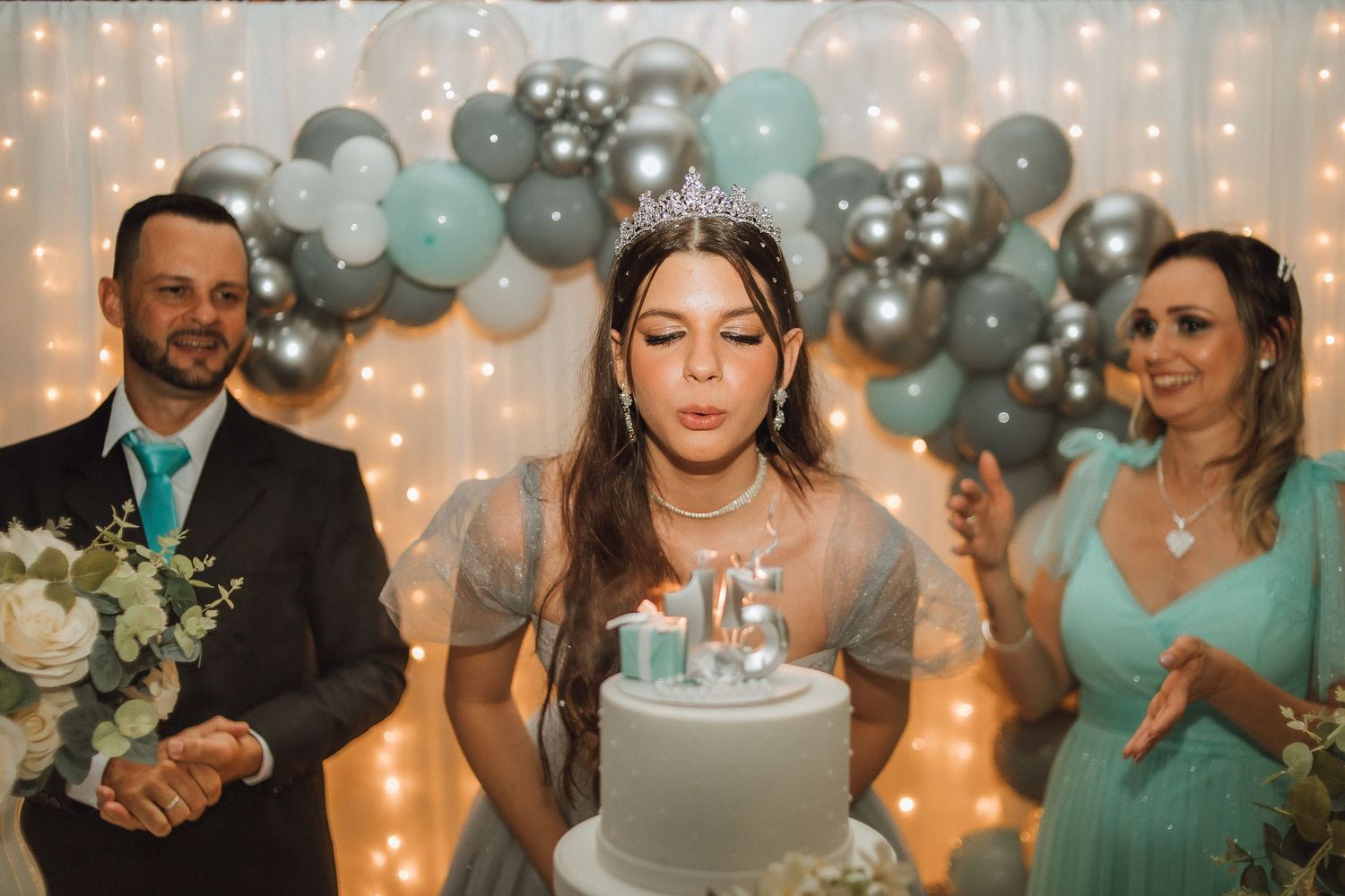 Teen girl blowing out candles on white birthday cake wearing tiara, flanked by parents at elegant quinceañera celebration.