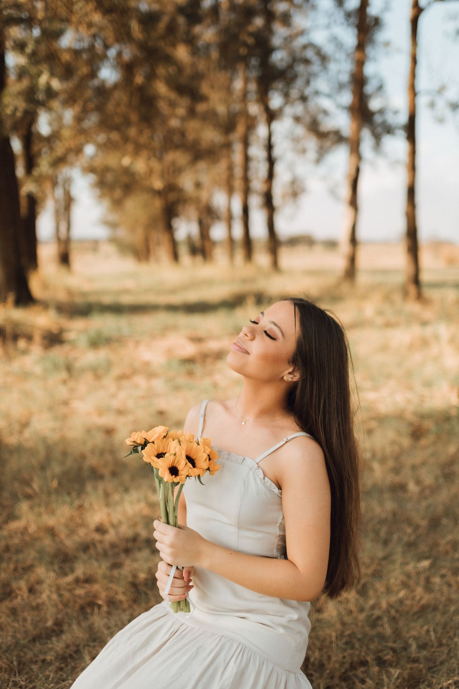 Young woman in white dress holding yellow sunflowers, sitting in a golden field with trees in the background.
