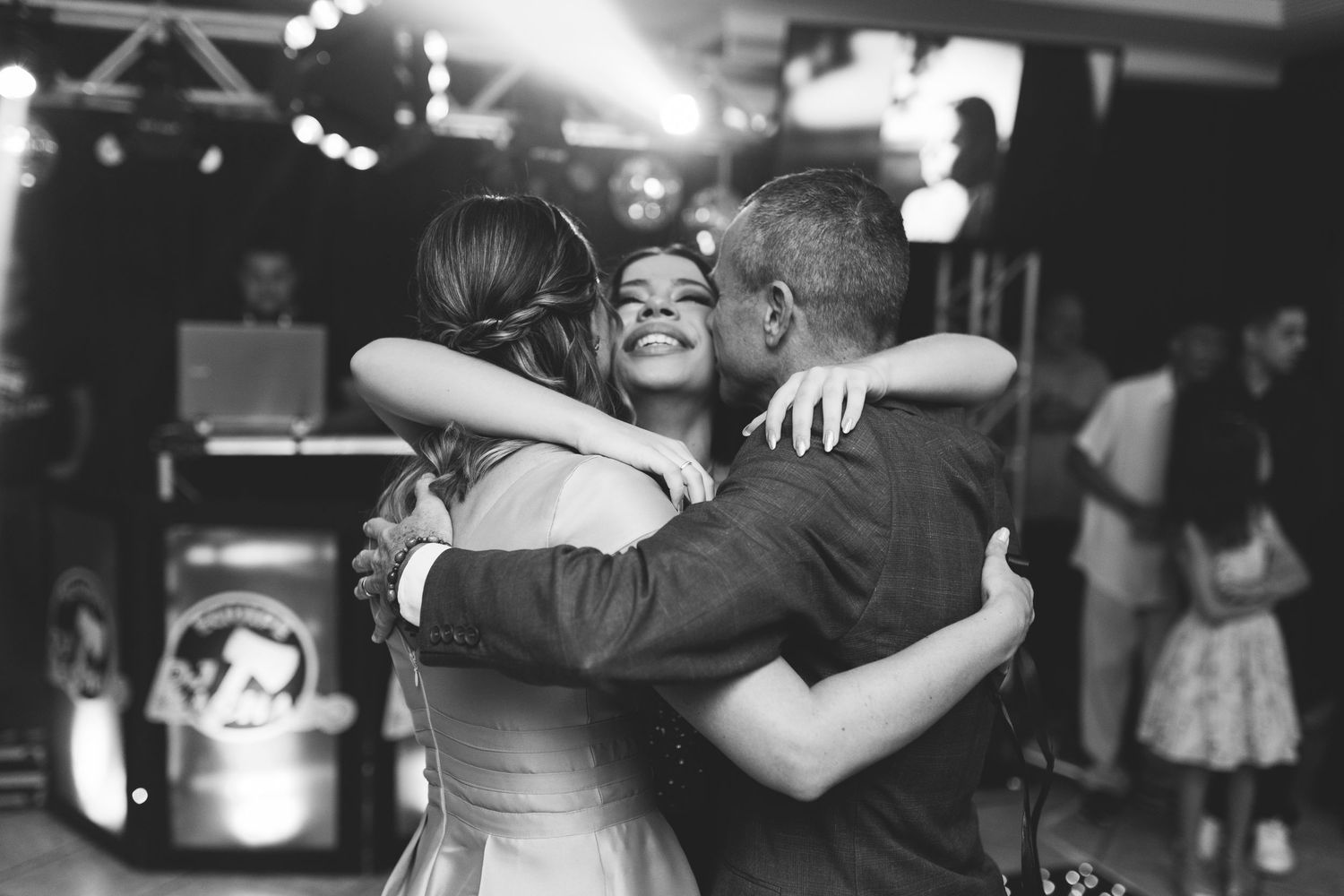 Black and white photo of three people sharing a warm group hug on a dance floor at a celebration.