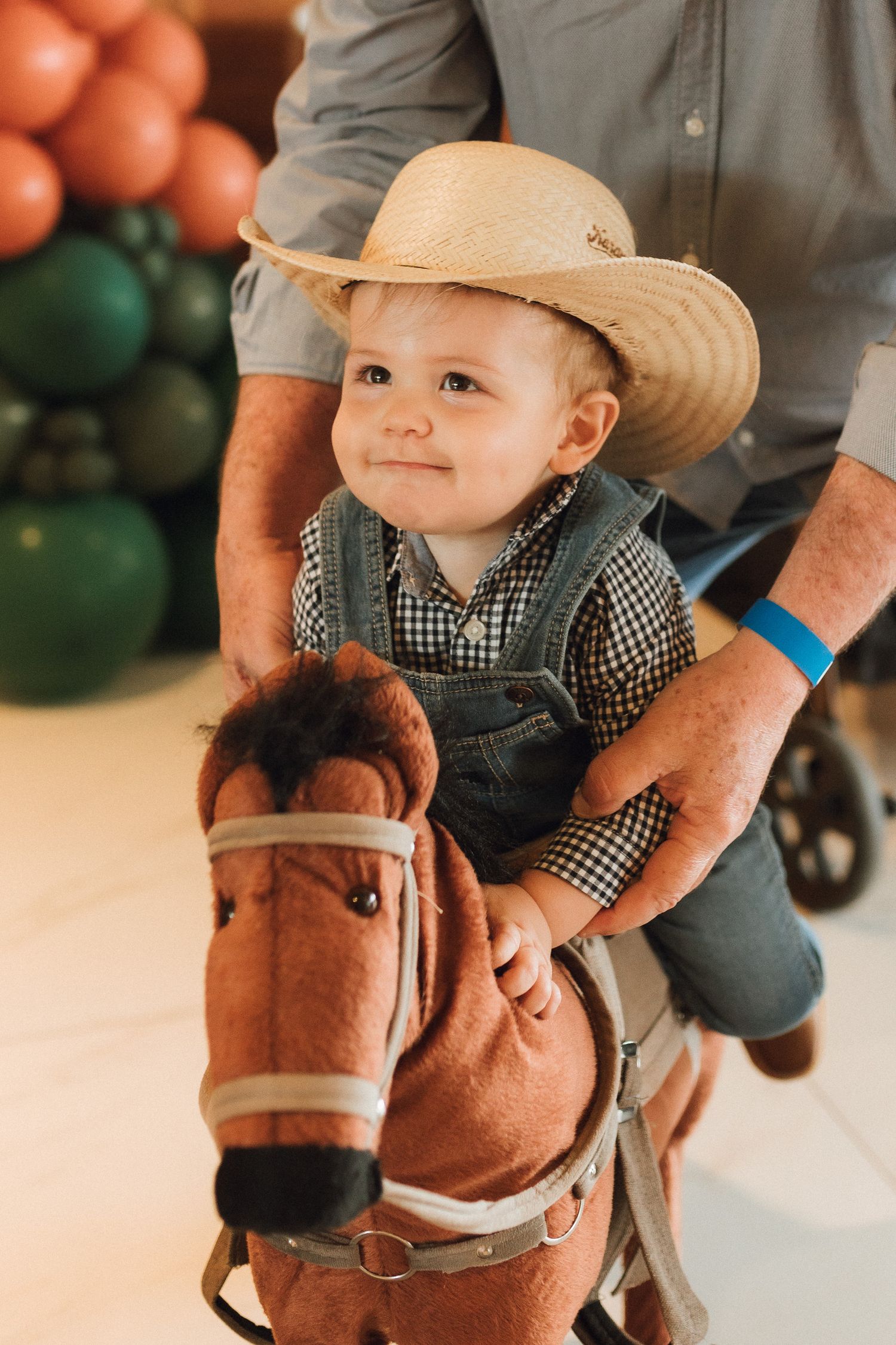 Smiling baby in cowboy hat and overalls sitting on a toy horse at a western-themed birthday party.