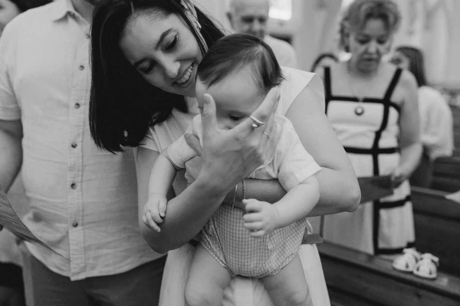 Black and white photo of a smiling woman holding a baby at a baptism ceremony inside a church.