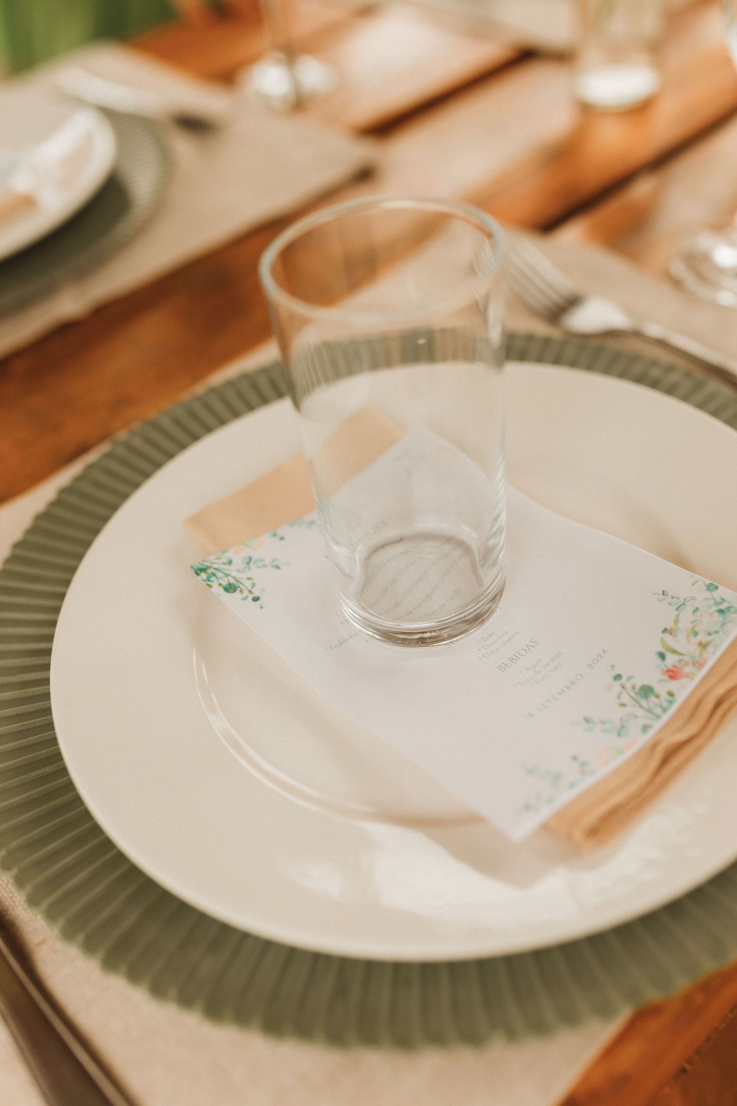 Elegant table setting with green charger plate, white dinner plate, floral menu card, and clear glass on wooden table.