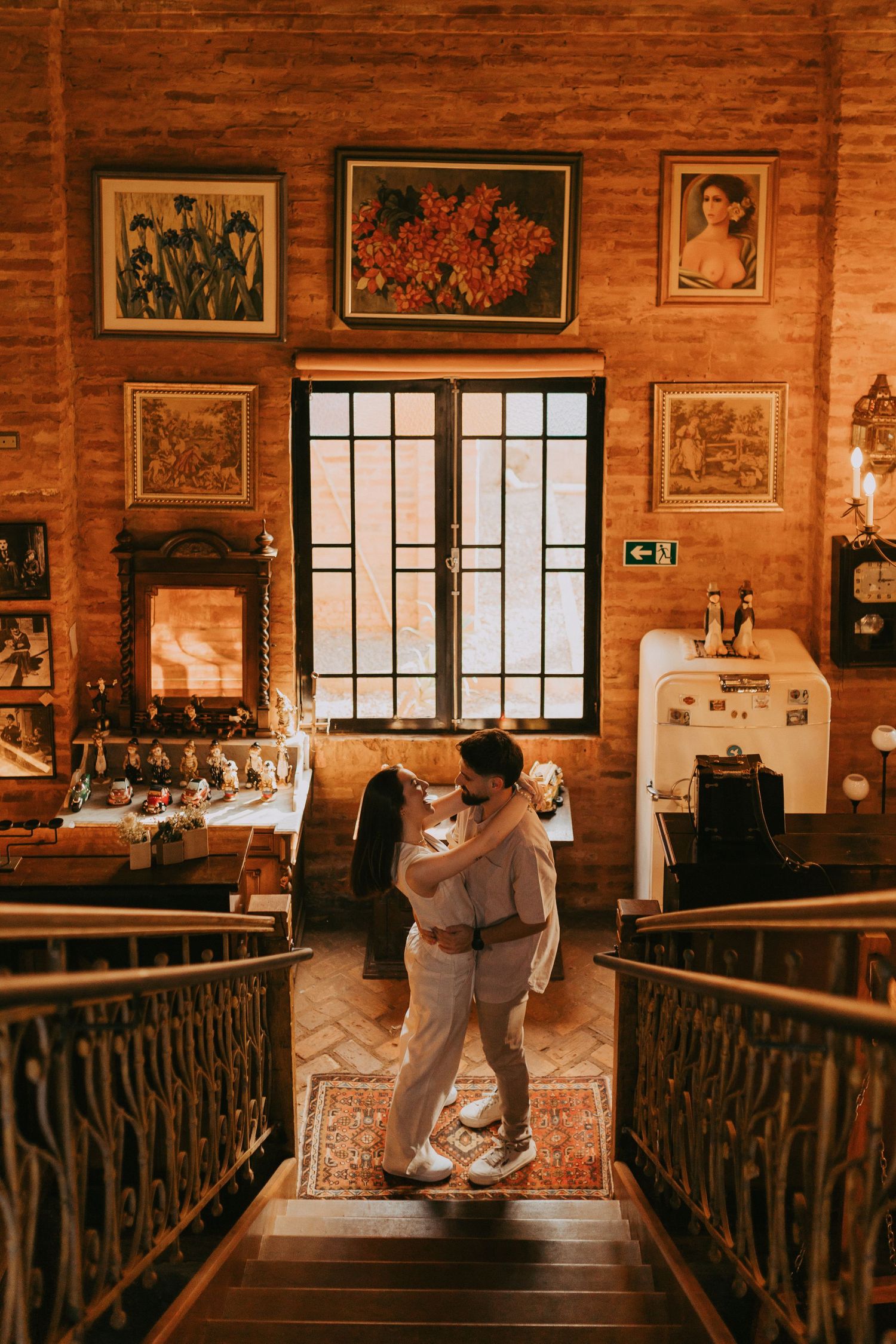 Couple embracing in a warmly lit rustic room with brick walls, framed artwork, and a patterned rug.