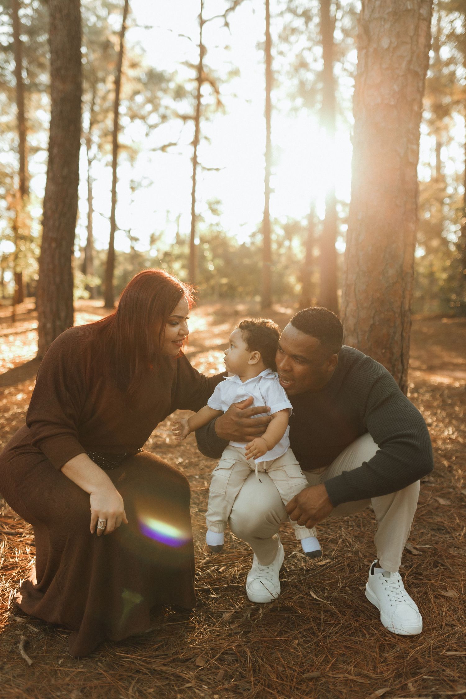 A joyful interracial family with a baby enjoying a sunny forest outing, surrounded by tall pine trees.