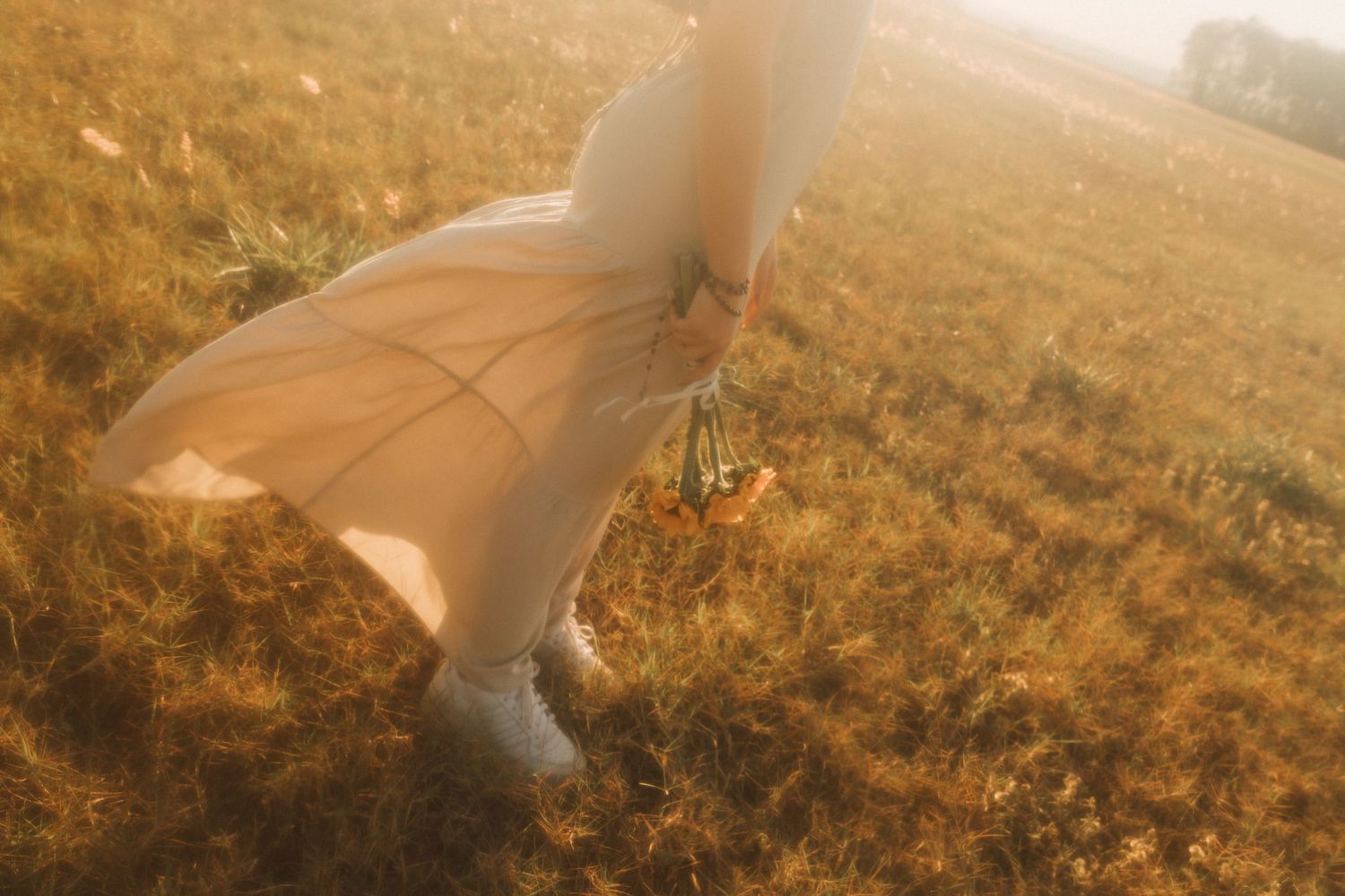 A woman in a flowing peach dress walks through a golden sunlit field, holding a bouquet of wildflowers.