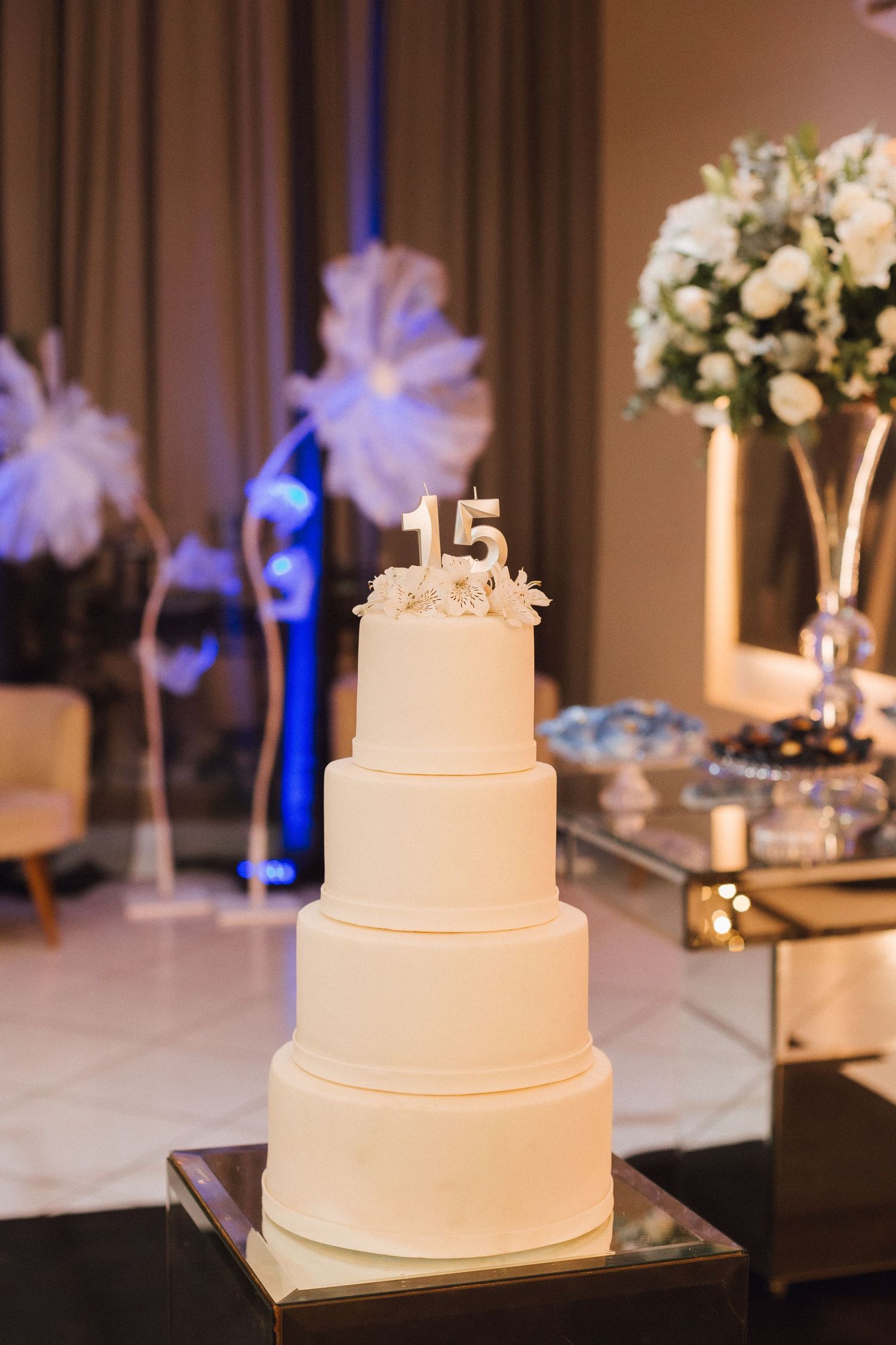 Elegant four-tier white quinceañera cake with number 15 topper displayed at a formal celebration venue.