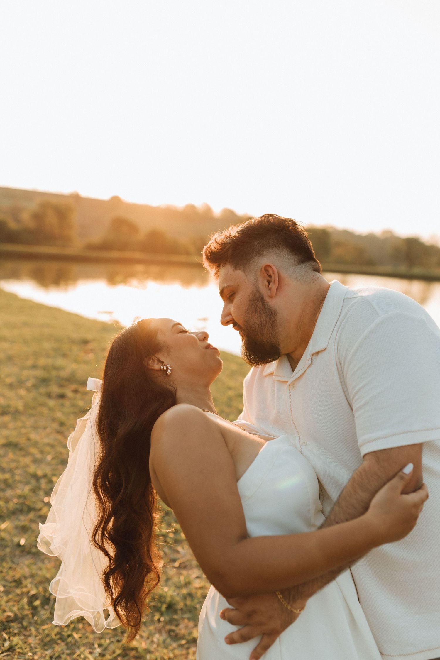 A couple in white attire shares a romantic moment at golden hour near a serene lake backdrop.