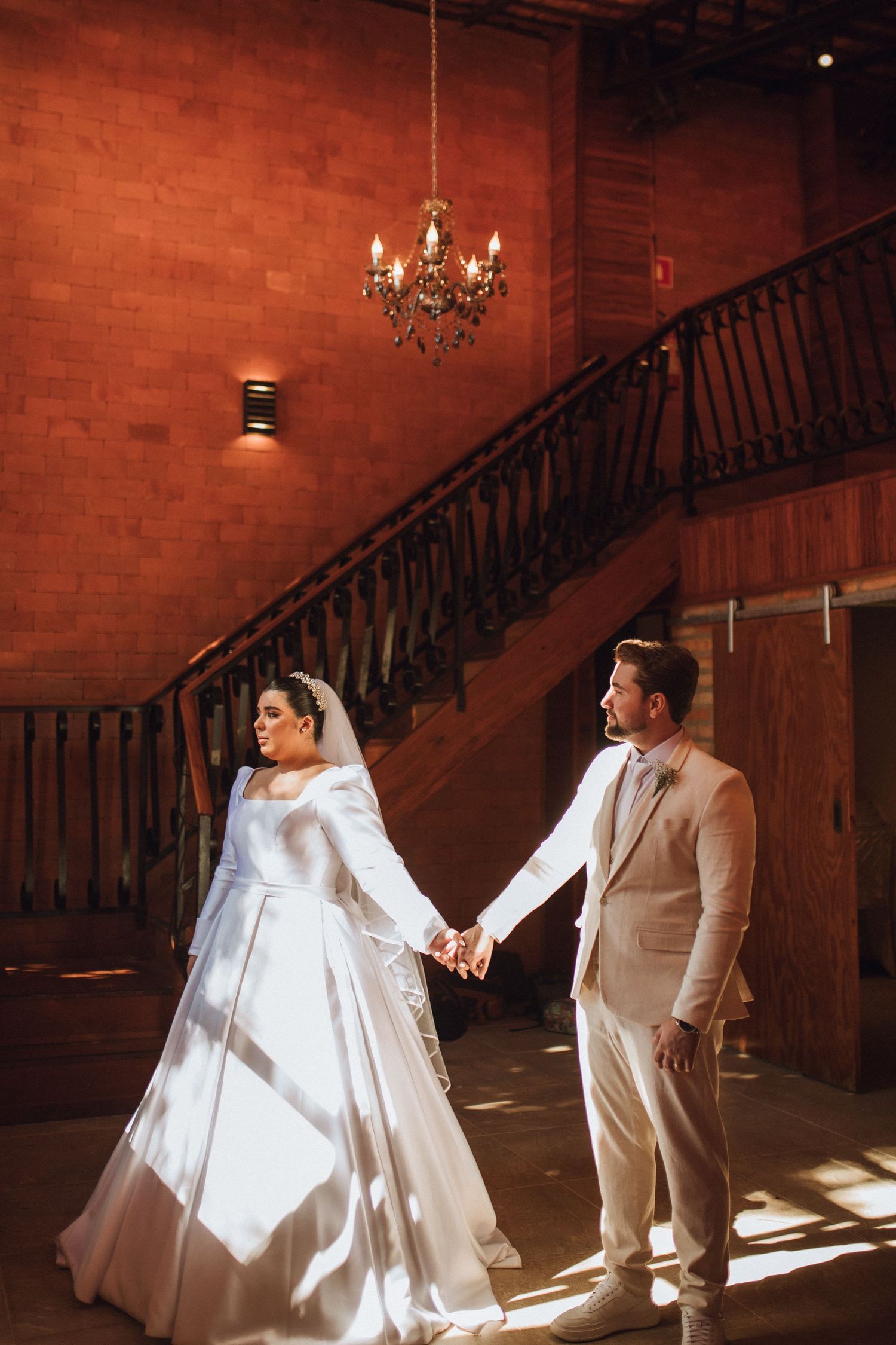 Bride and groom hold hands beneath a chandelier near a grand wooden staircase.