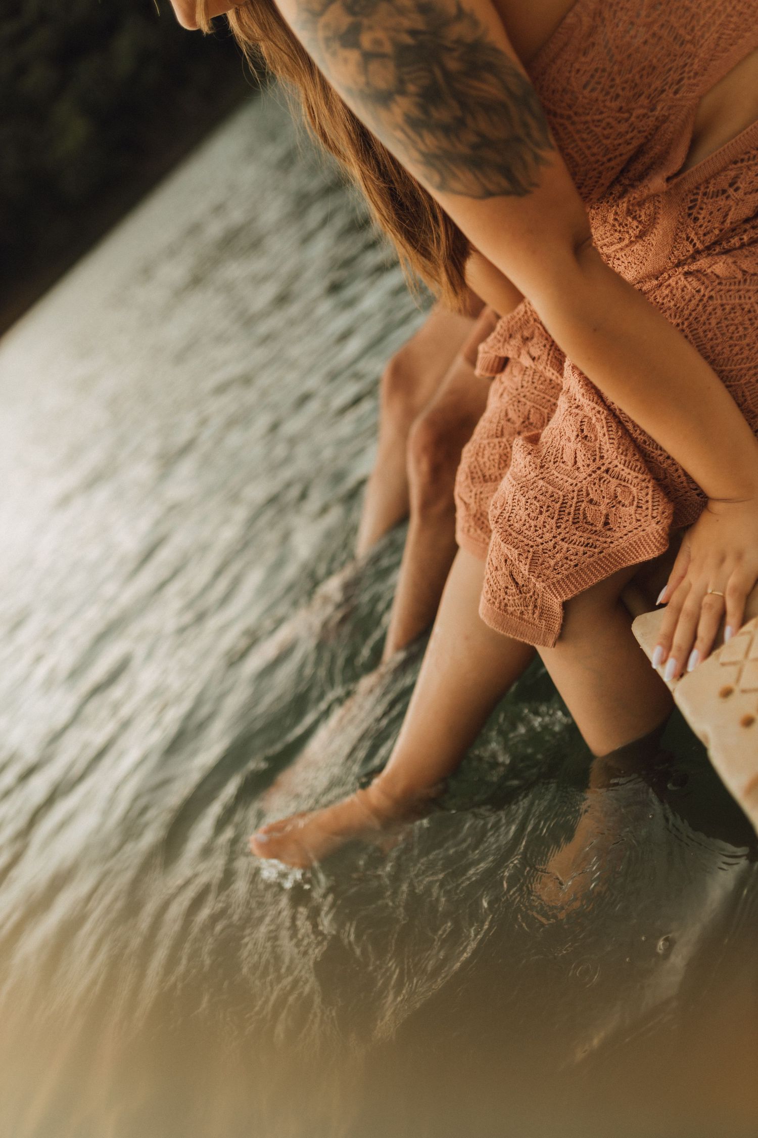 Woman in pink lace dress sitting on dock with feet in water.