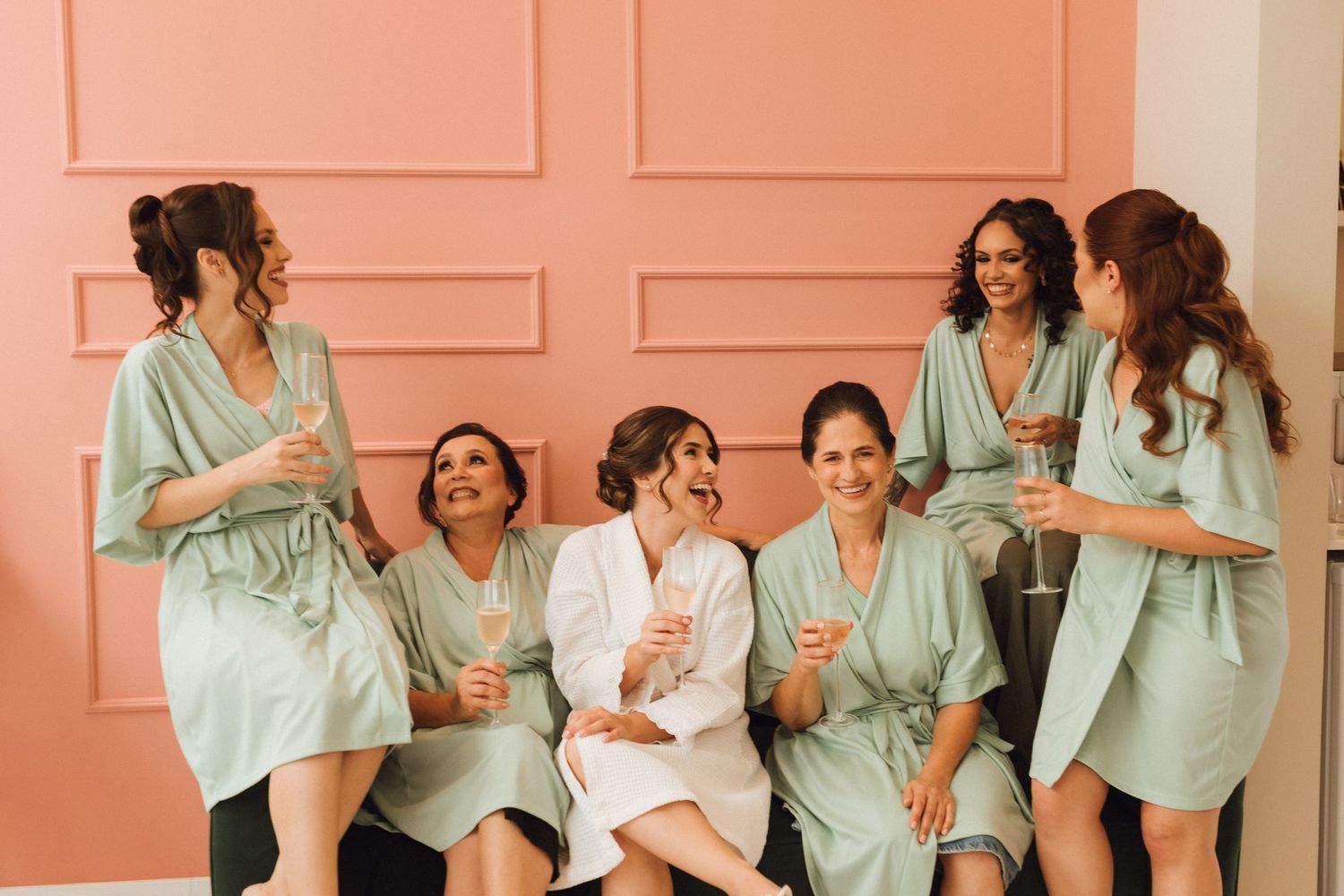 Bridesmaids in sage green robes laughing and celebrating with champagne against a pink wall.