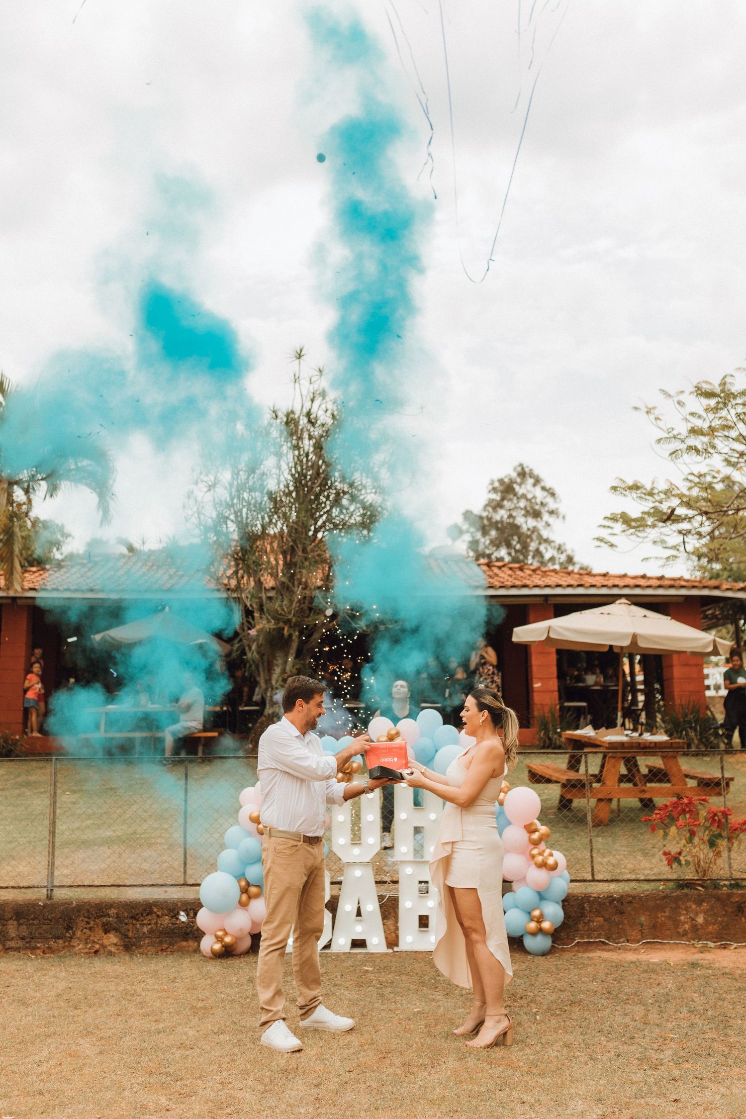 Couple releasing blue smoke bomb at outdoor gender reveal party revealing baby boy in front of OH BABY balloon display.