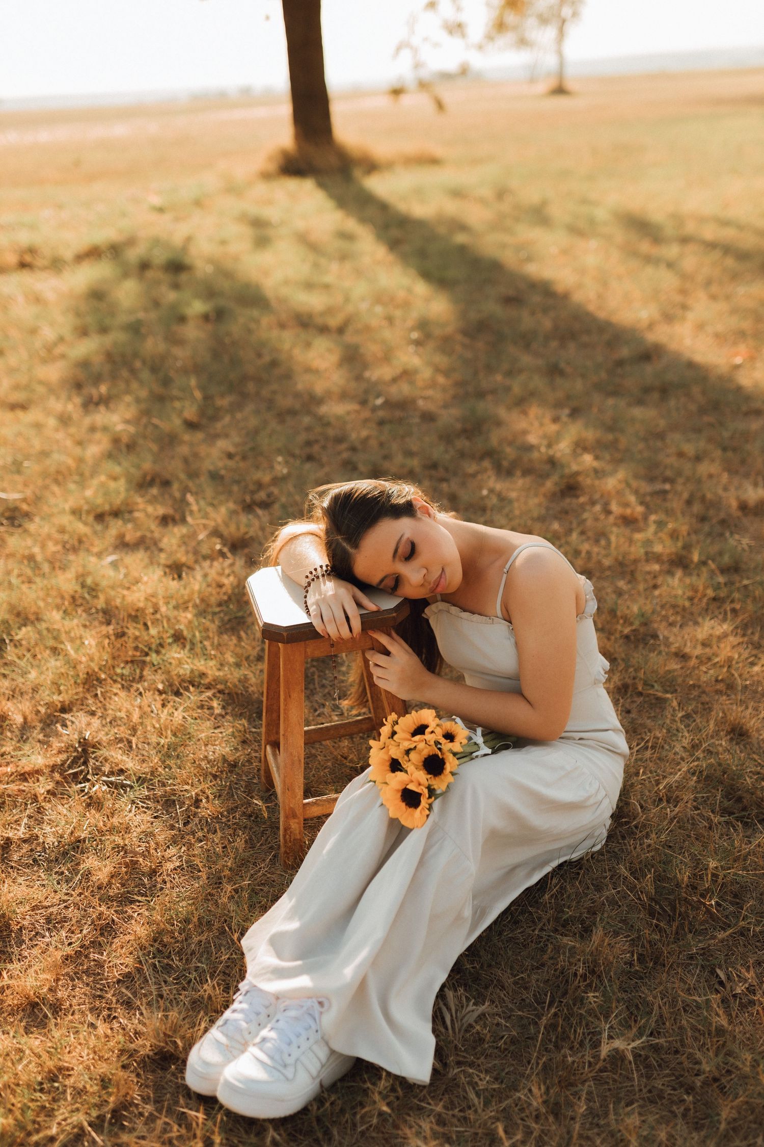 Young woman in white dress sitting on grass holding sunflowers, leaning on wooden stool at golden sunset.
