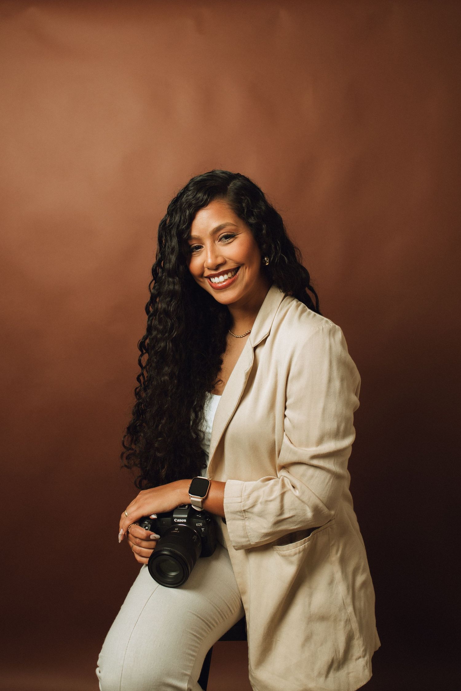 Smiling female photographer holding a camera poses against a warm brown studio background.