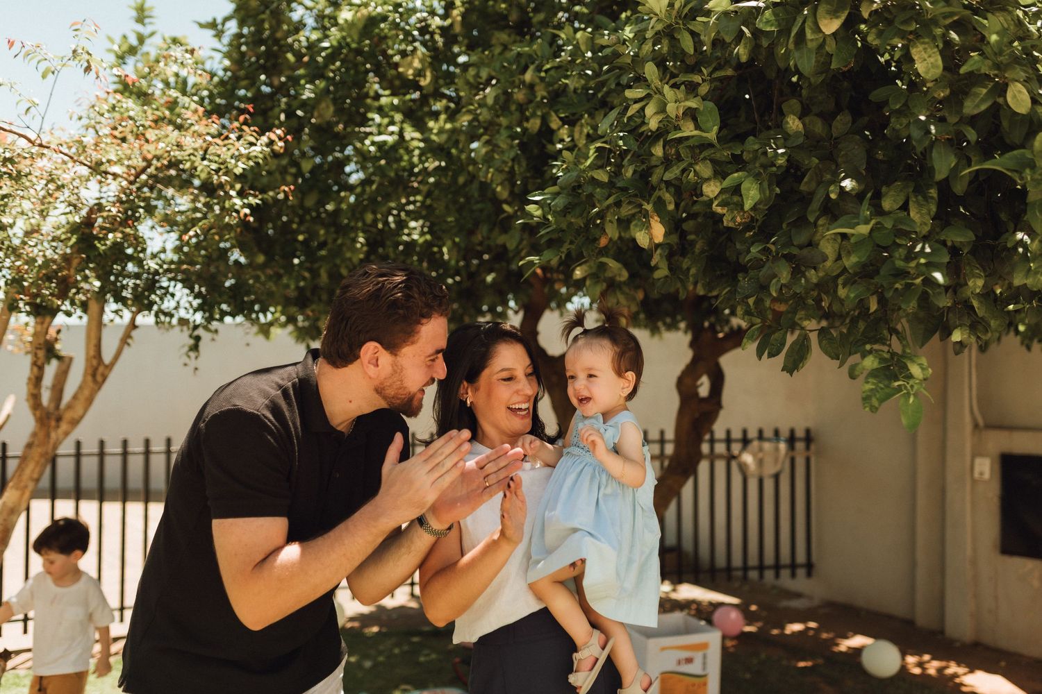 Happy family with parents clapping and holding toddler outdoors under green tree on sunny day.
