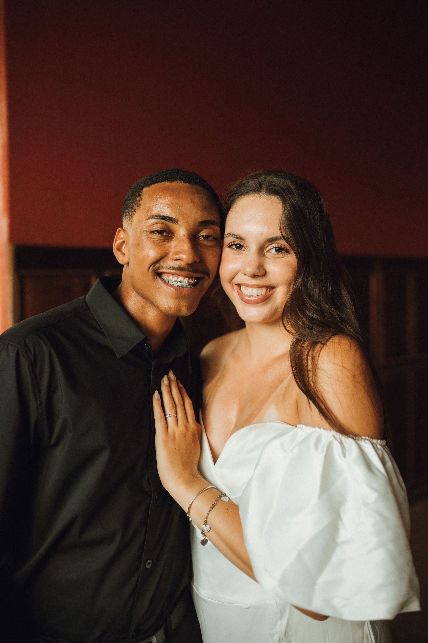 A smiling interracial couple poses together, man in black shirt and woman in white off-shoulder dress, against a dark red wall.