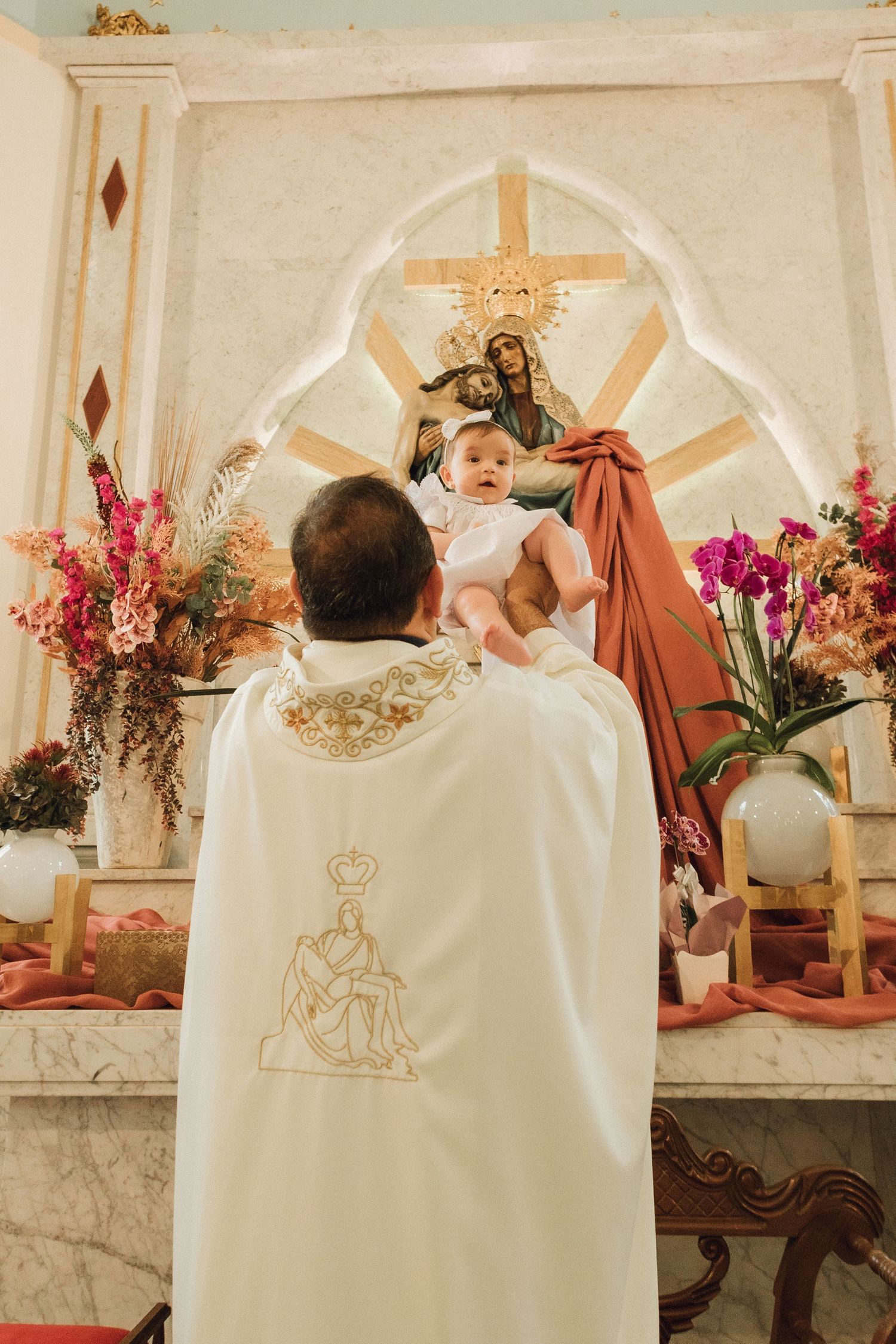 Priest lifting baby toward Virgin Mary altar during Catholic baptism ceremony with floral arrangements.